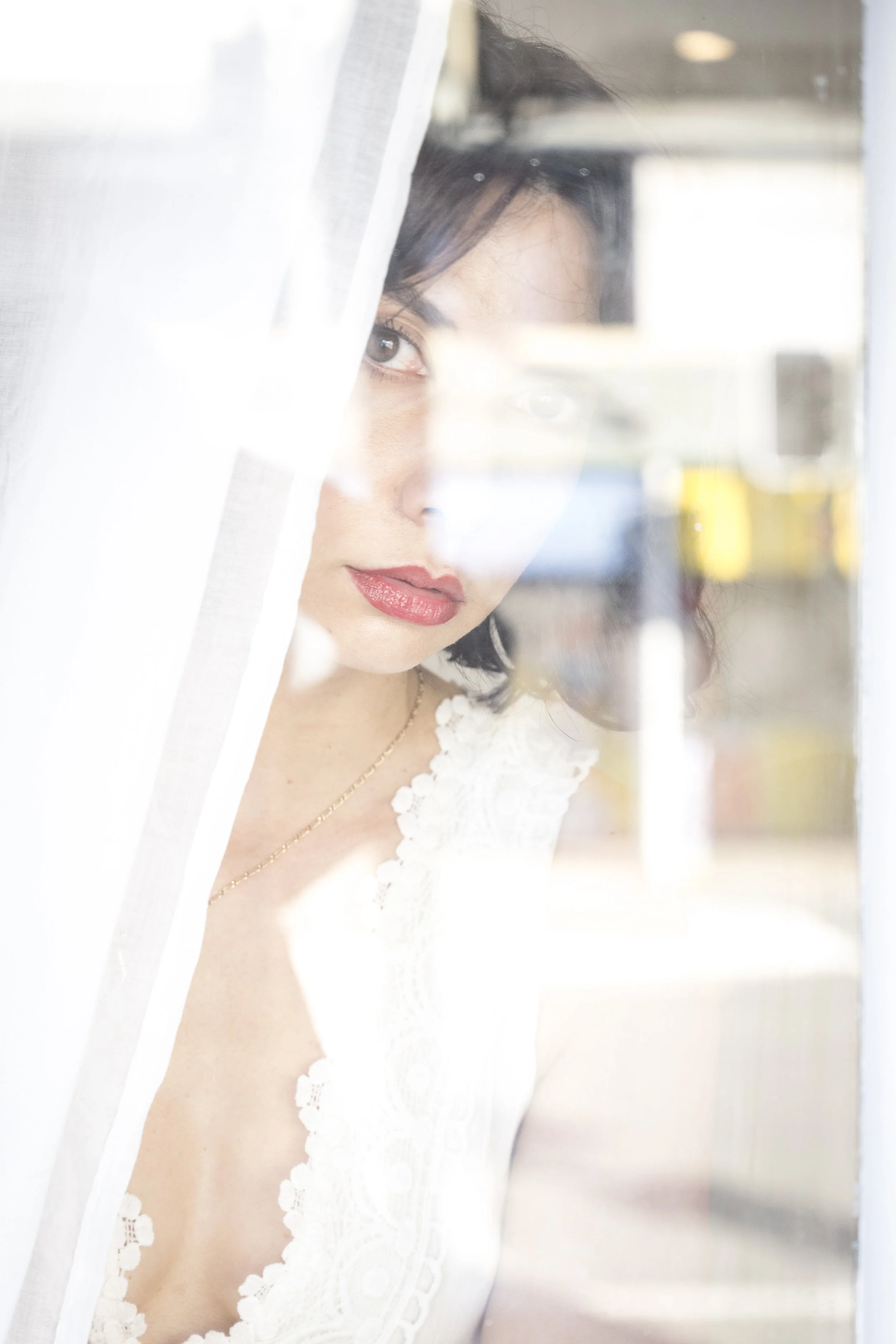 A woman with dark hair, red lipstick, and a white lace top looks through a window, partially obscured by a sheer curtain, with a blurred background.