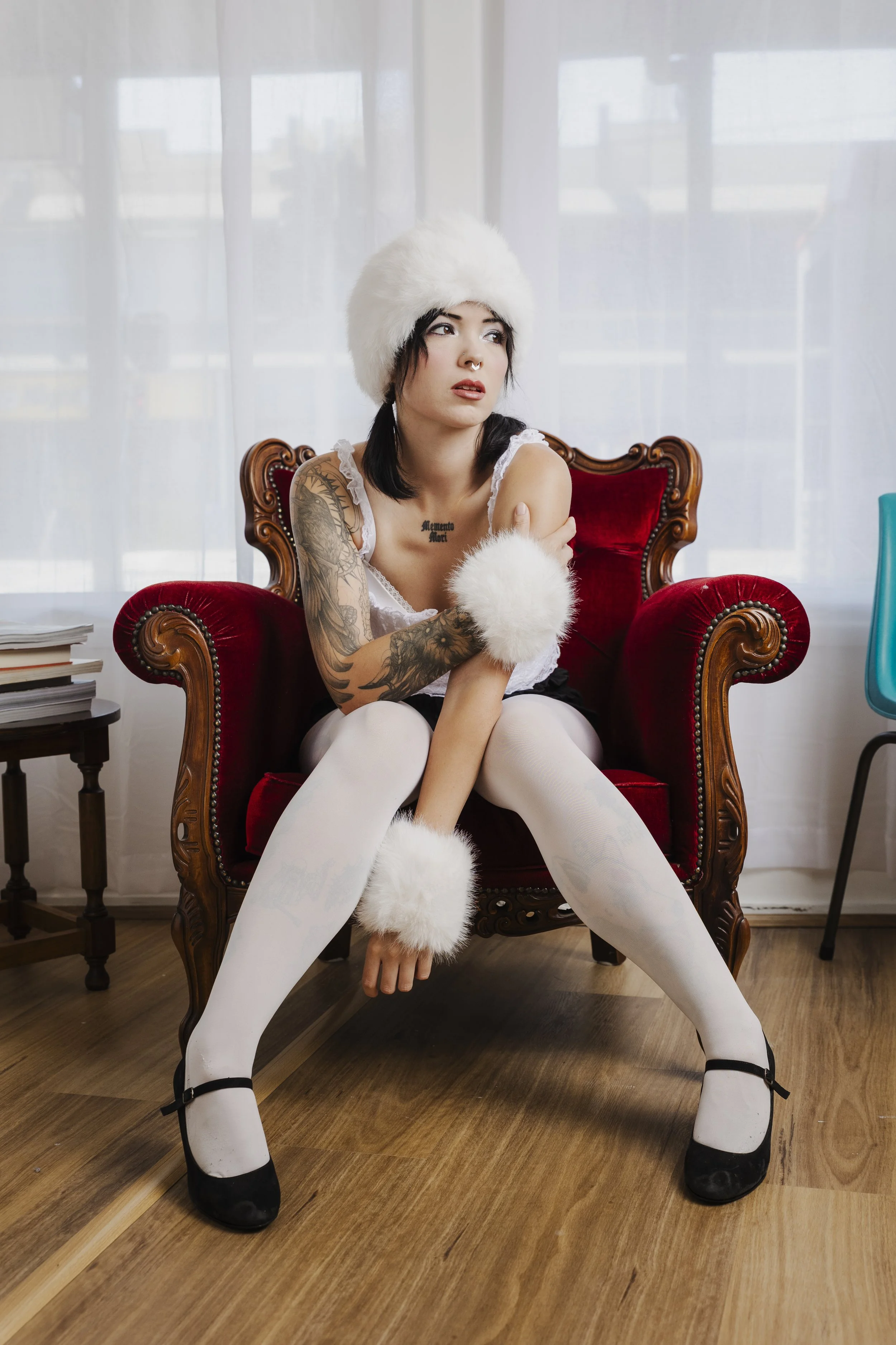 A woman sitting on a vintage red armchair in a room with white curtains, wearing white stockings, black shoes, and fluffy white accessories, with tattoos visible on her arms and chest.