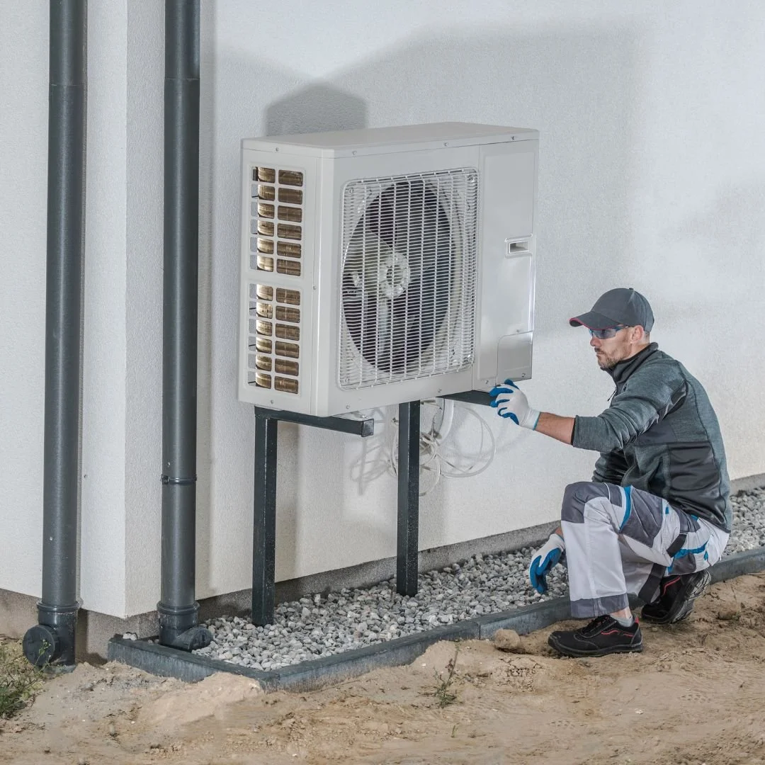 Technician inspecting an outdoor air conditioning unit mounted on a stand beside the wall of a building.