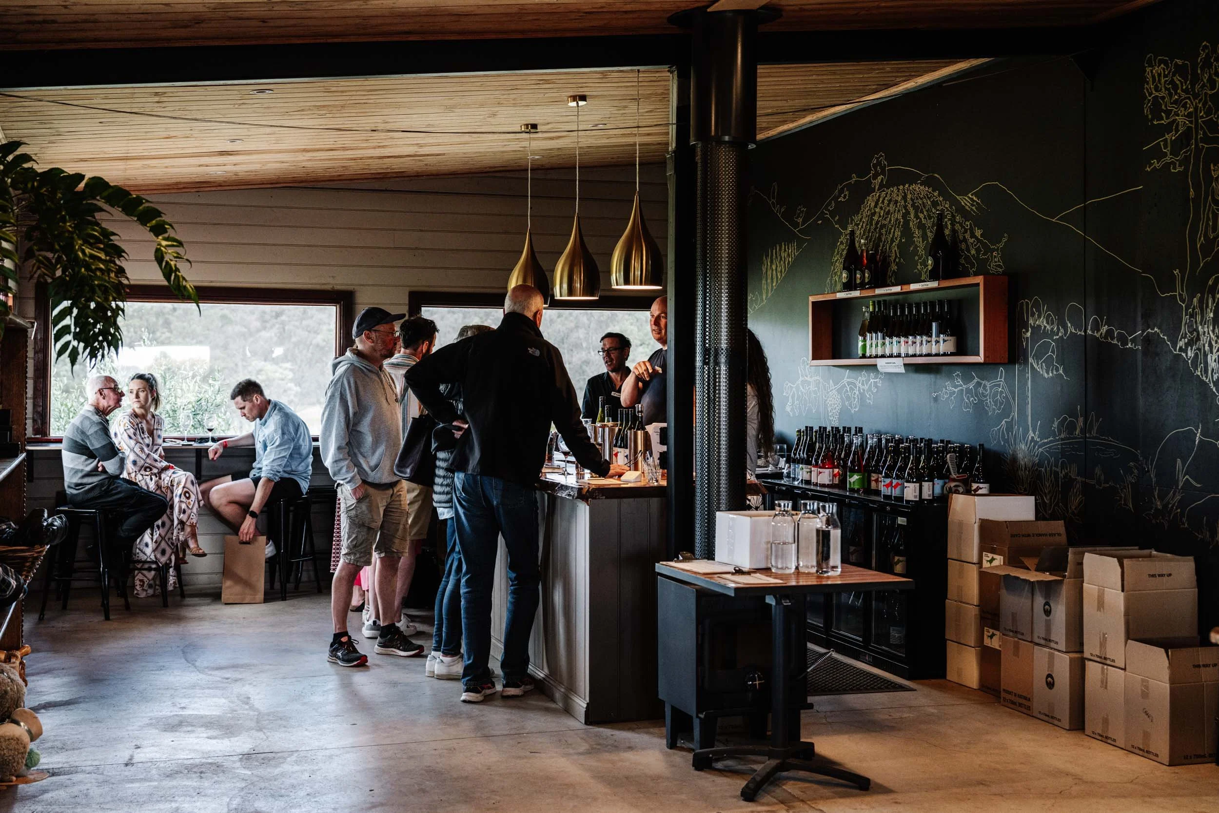 A group of people gathered around a bar counter in a cozy . Some are chatting, others are ordering drinks. The cafe has large windows and a decorative chalk wall with a mountain landscape drawing. There are hanging pendant lights and cardboard boxes stacked in the corner.