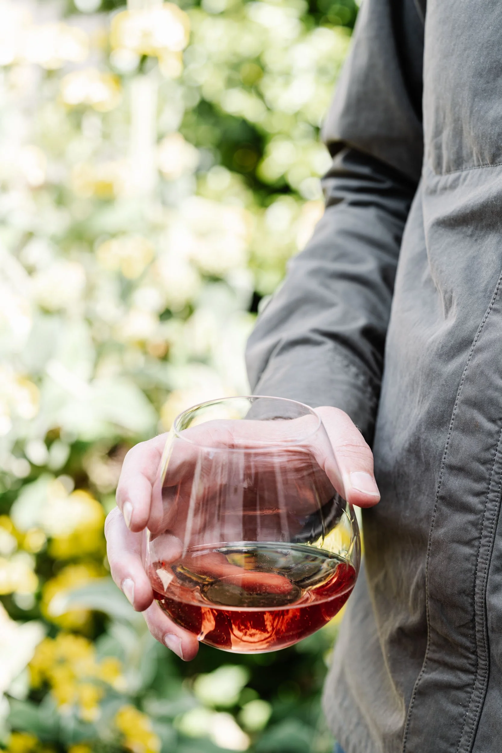 Person holding a glass of rosé wine outdoors with greenery in the background.