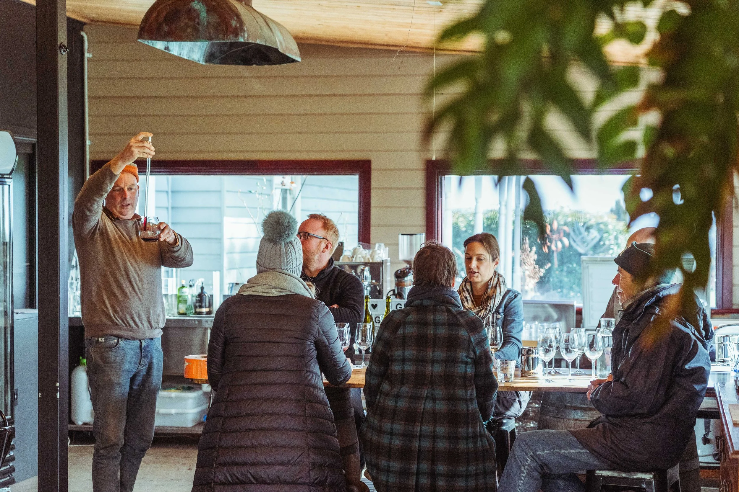 A group of people in a cozy indoor setting, participating in a wine tasting with a man pouring wine from a pipette, while others sit at a table with wine glasses.