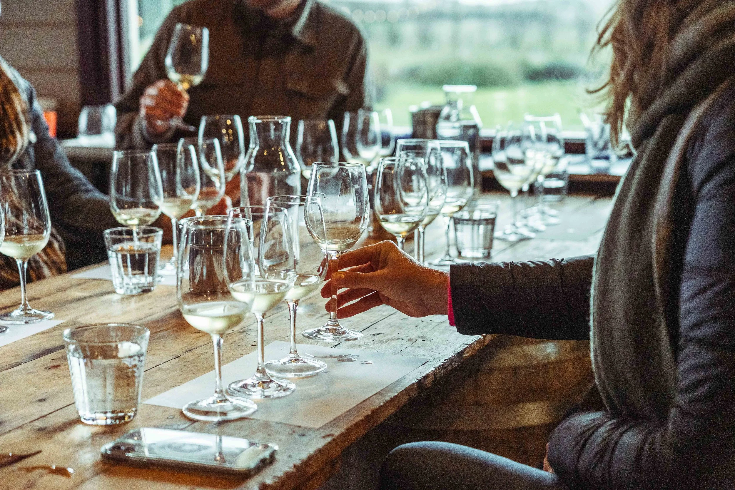 People participating in a wine tasting, tasting white wine in glasses on a rustic wooden table with water glasses and a phone, in a room with large windows showing an outdoor view.