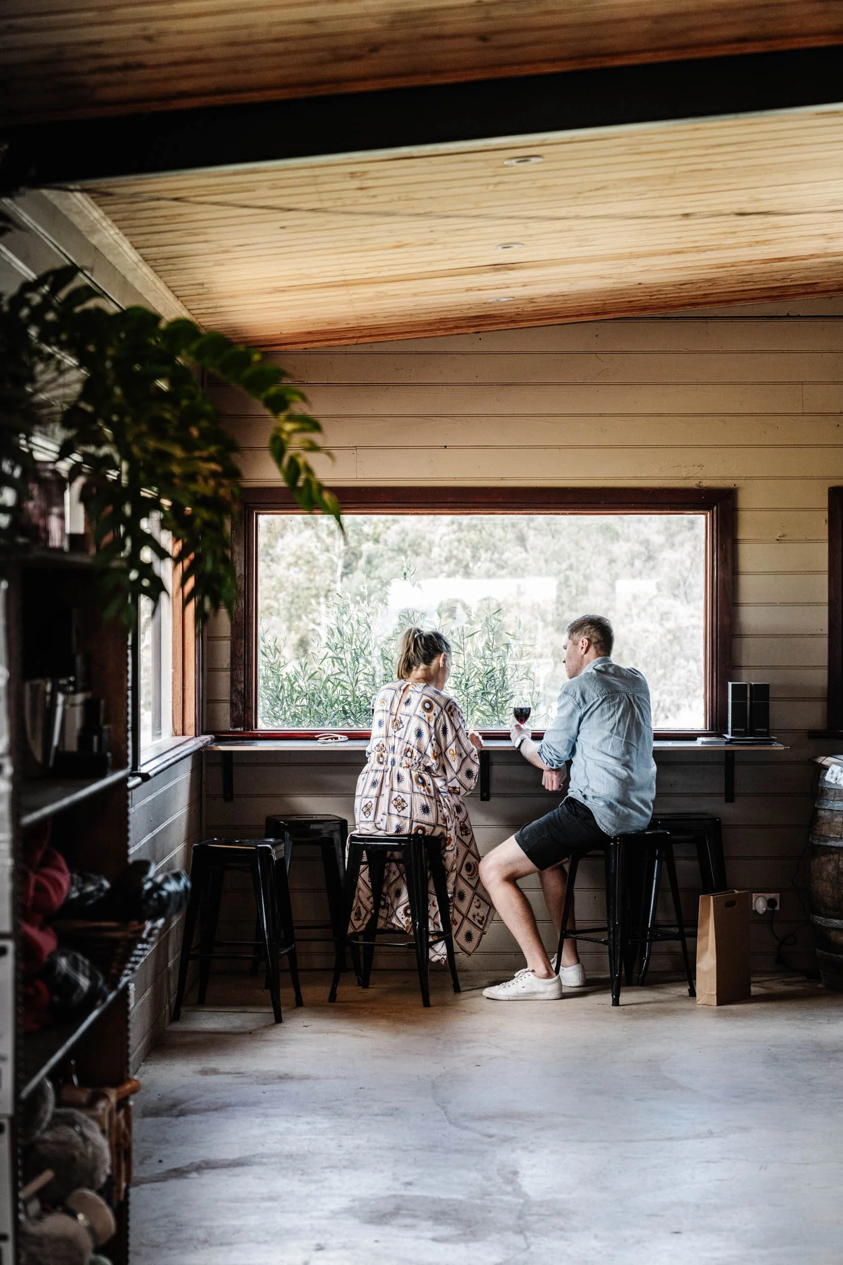 A man and woman sitting on bar stools by a large window, with a glass of red wine, in a cozy room with wooden walls.