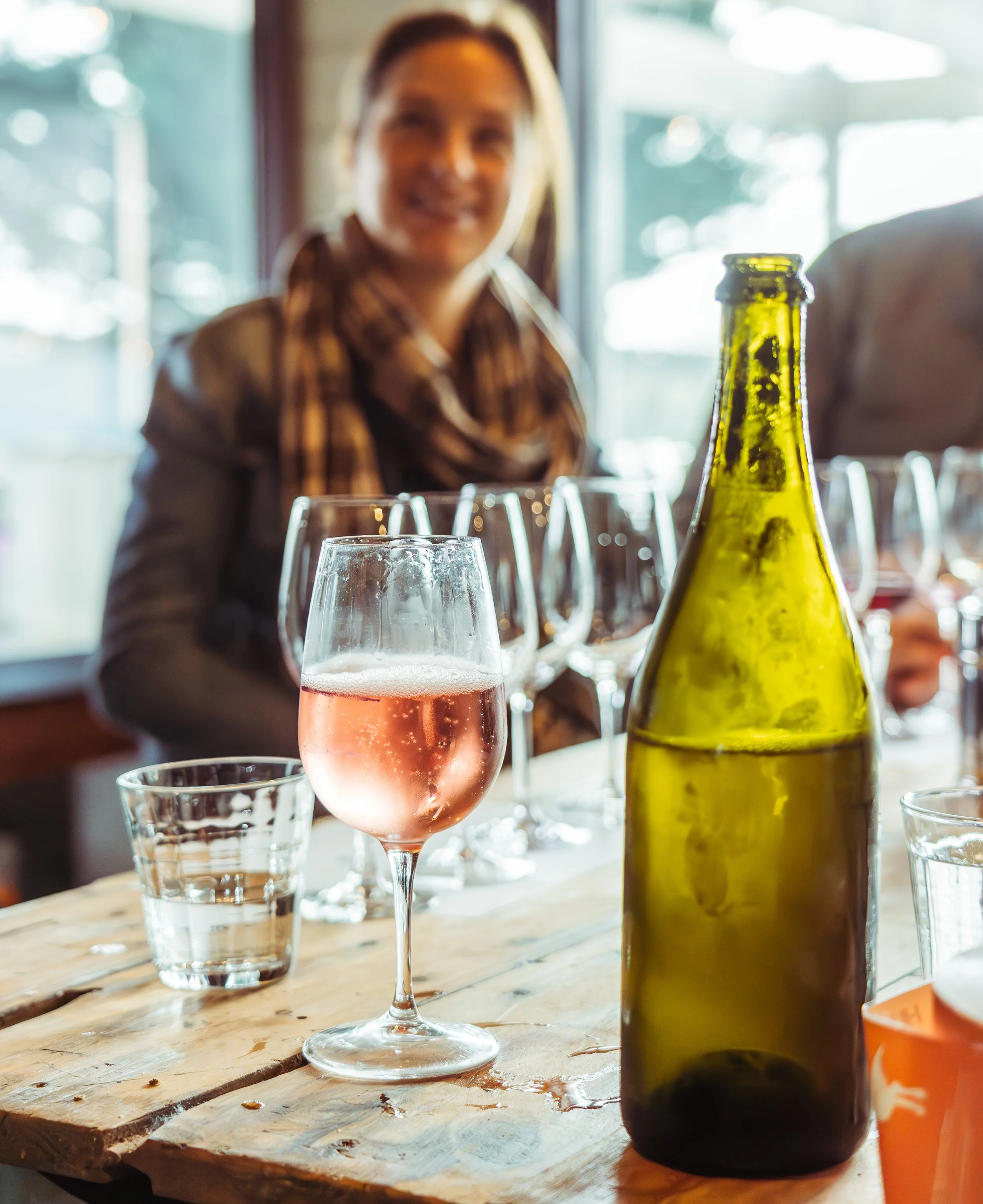 A woman smiling at a wine tasting with a glass of rosé wine, a green wine bottle, and several empty wine glasses on a wooden table.