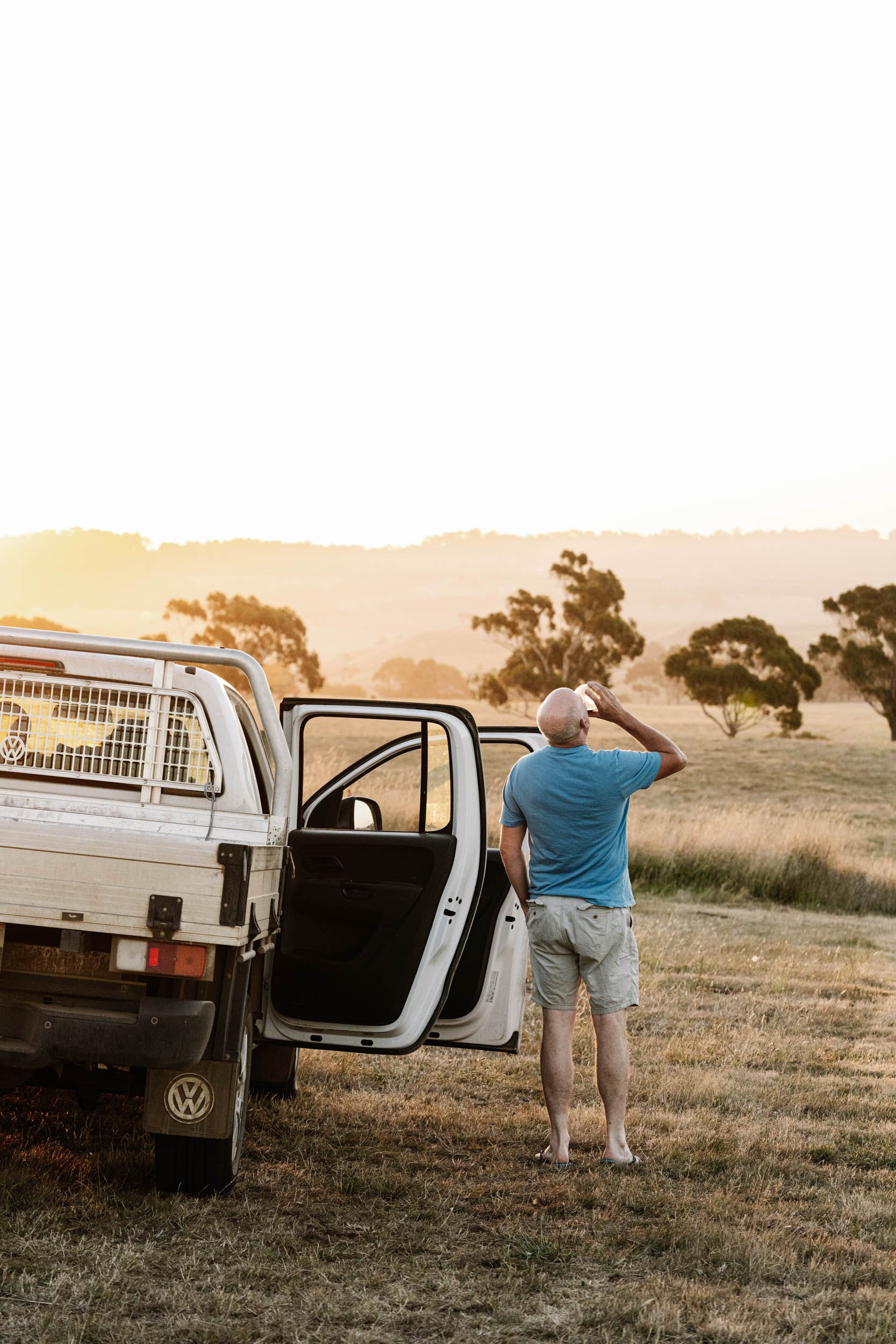 A man standing barefoot outdoors beside a white pickup truck during sunset, looking into the distance and drinking wine.