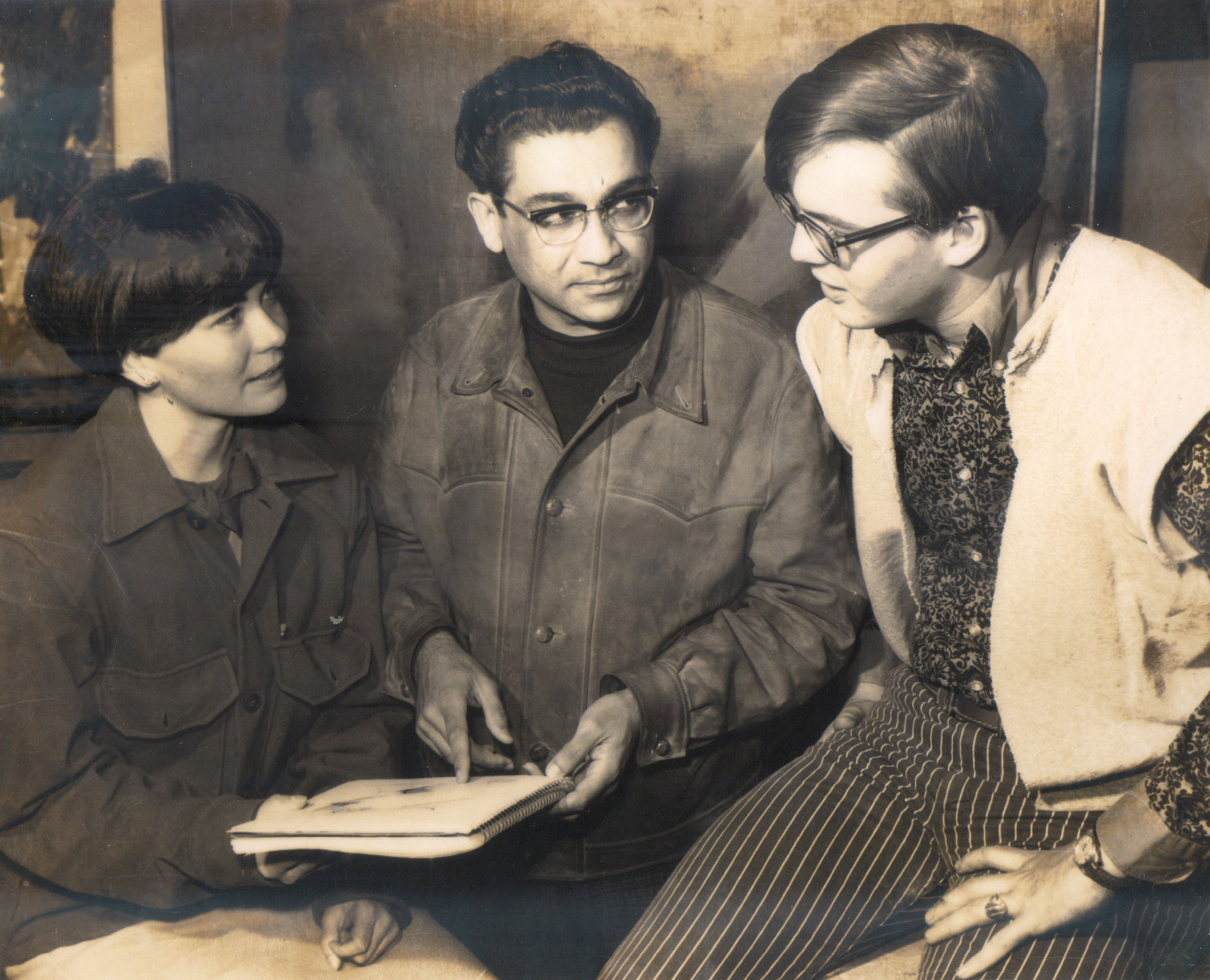 Black and white photo of three people, two men and one woman, engaged in a discussion while looking at a notebook. The woman is wearing a jacket and has short hair. The man in the middle is wearing glasses and a leather jacket, holding the notebook. The man on the right is wearing glasses, a patterned shirt, and a sweater vest, with striped pants.