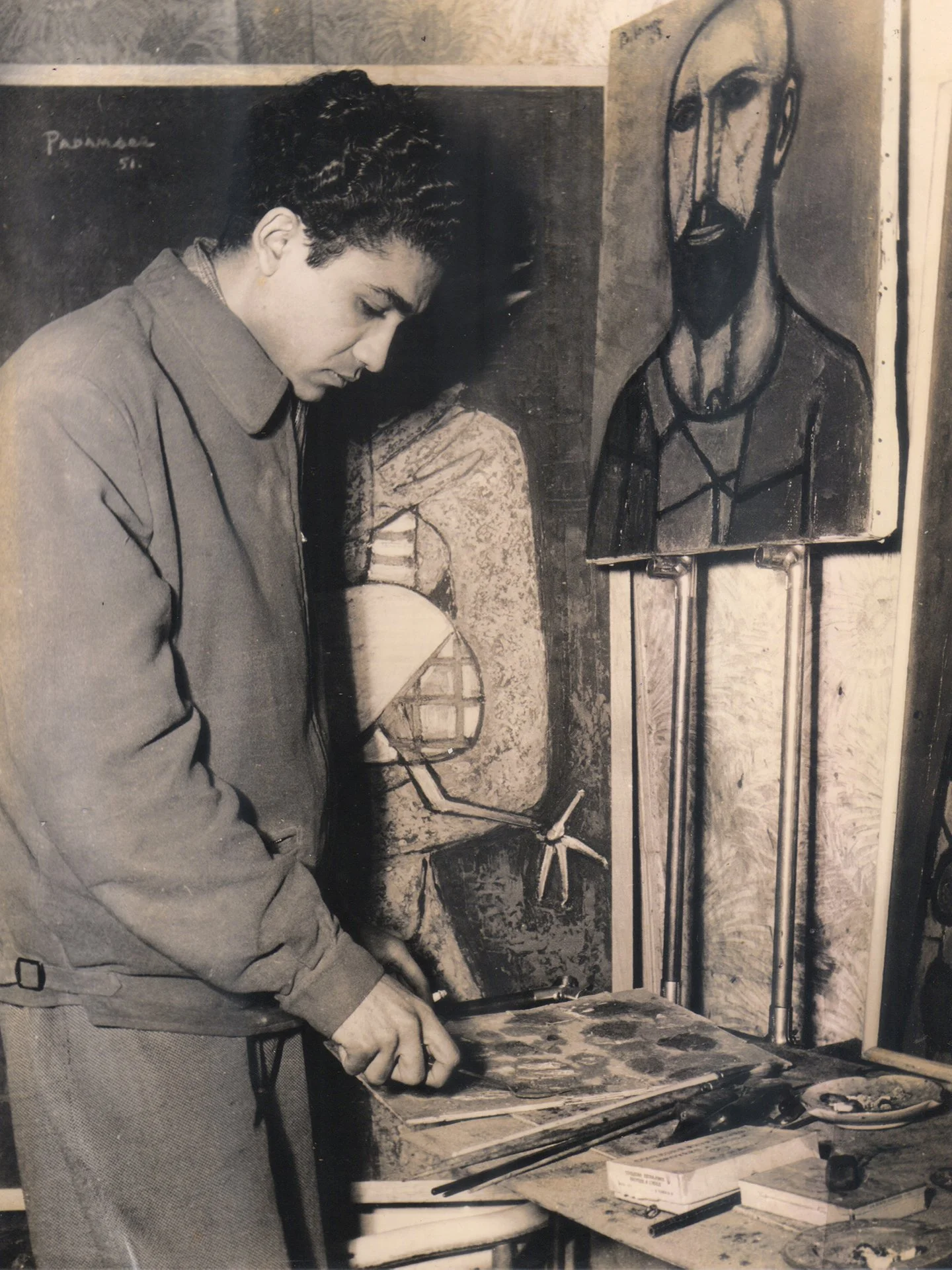 A man in a jacket and pants standing at a worktable, painting or working on a canvas in a studio. Behind him, there are several artworks, including a portrait of a man with a beard and an abstract piece with a pie chart and windmill, all in black and white.