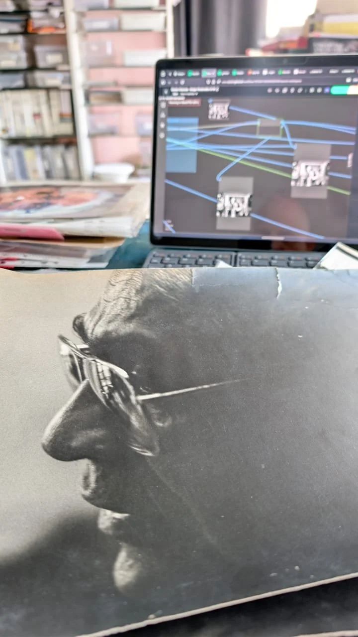 Close-up of a black and white photograph of a person wearing sunglasses on a book or magazine cover, with a blurred laptop showing a digital design on the screen in the background.
