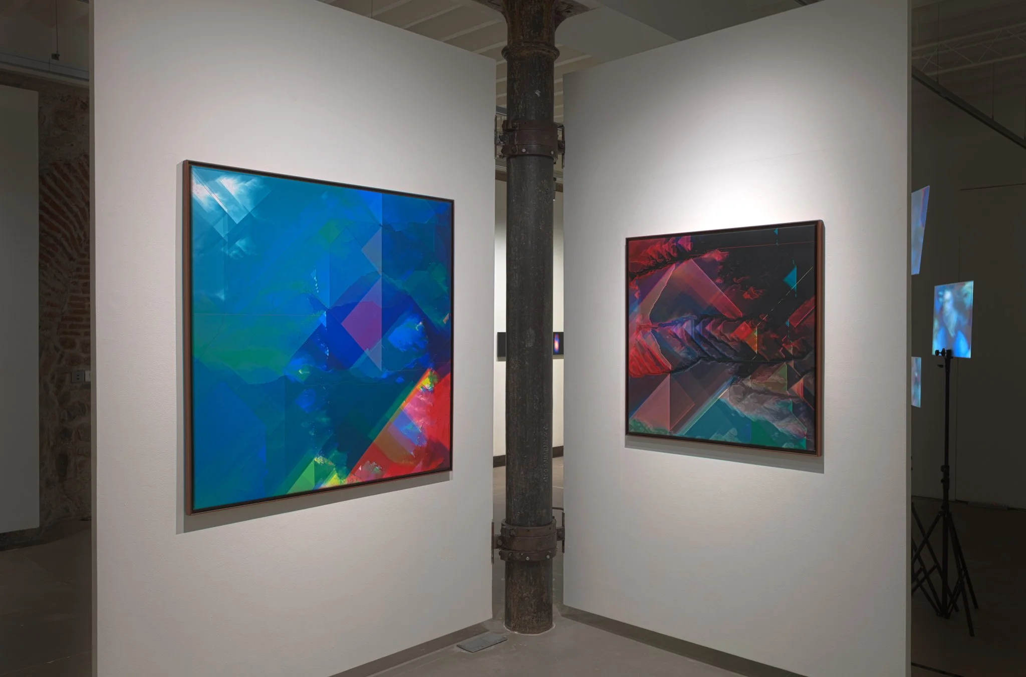 Two abstract digital artworks hanging on white gallery walls, separated by a black metal support column.