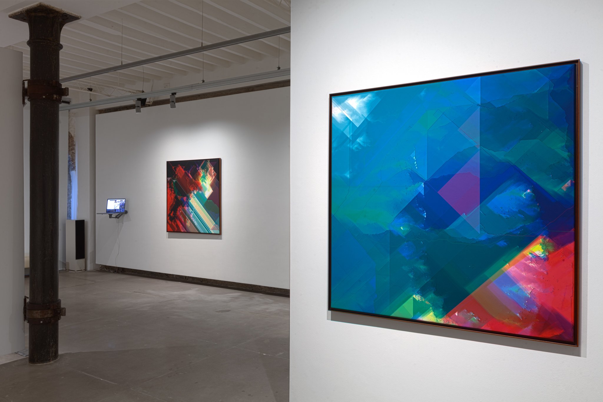 View of an art gallery with abstract digital paintings, one large on the right wall in shades of blue and purple, and a smaller colorful piece on the back wall, alongside a mounted digital display.