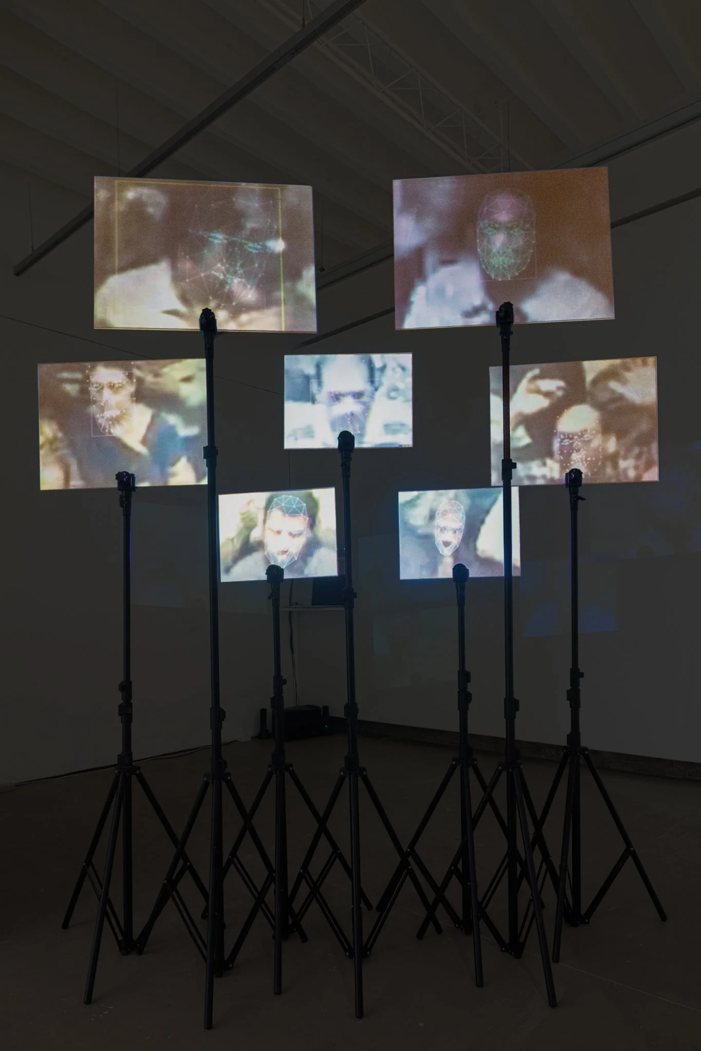 Multiple screens featuring blurred faces and digital graphics, mounted on adjustable tripods, in a dark gallery space. Digital Art