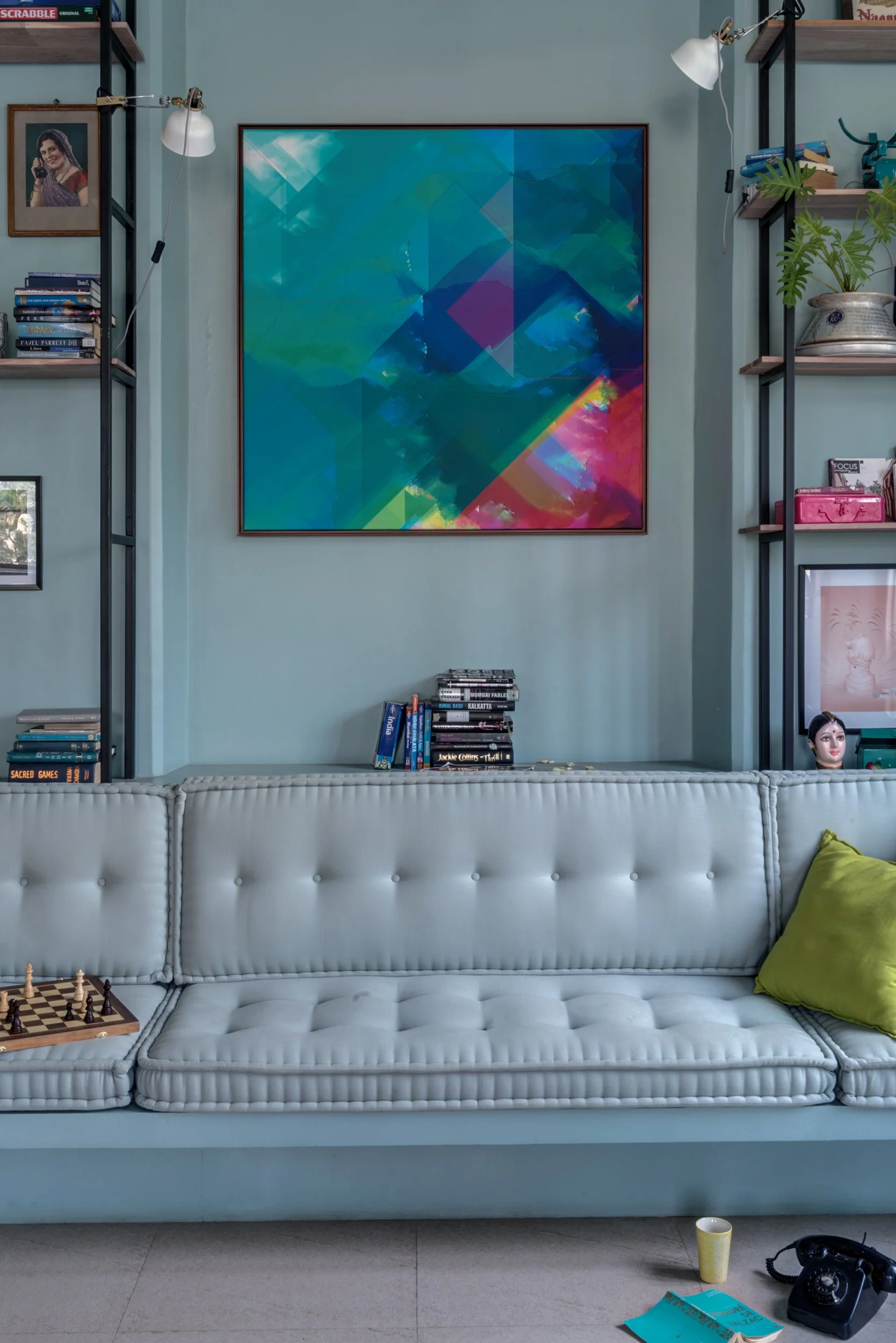 A living room with a light blue tufted sofa, a green pillow, a chessboard, and a small yellow cup on the floor. Behind the sofa, there are two black shelves filled with books, framed pictures, and decorative items. A large abstract colorful painting 