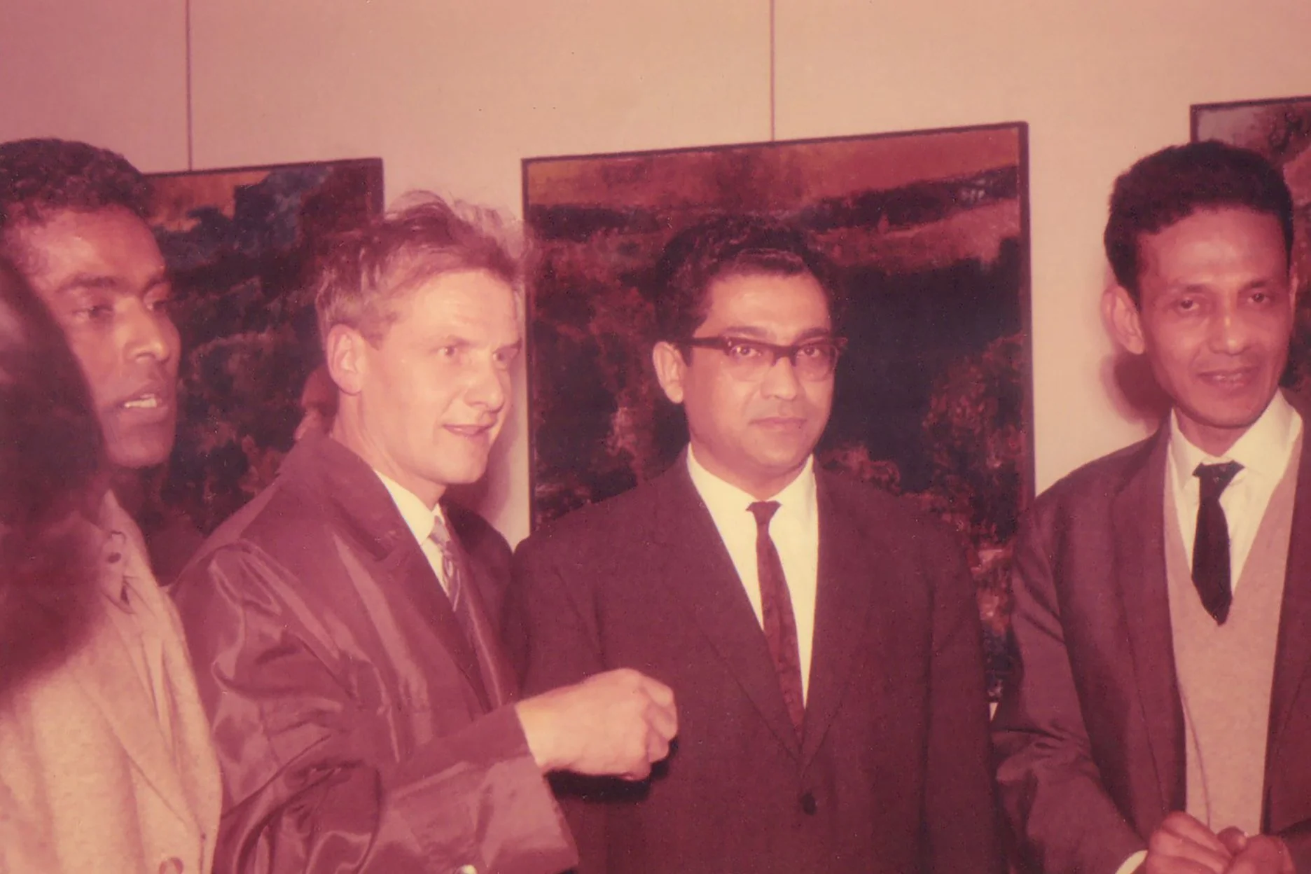 Four men in suits standing together at an art gallery with landscape paintings in the background.