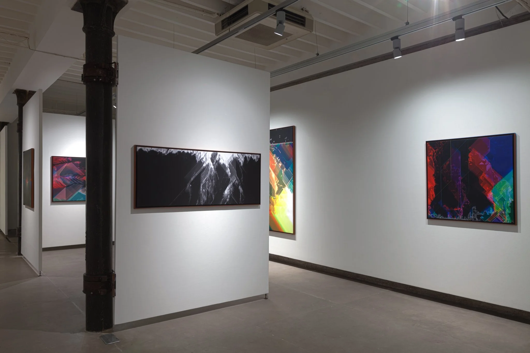 An art gallery with abstract and digital artwork displayed on white walls, with track lighting and an industrial black pillar.