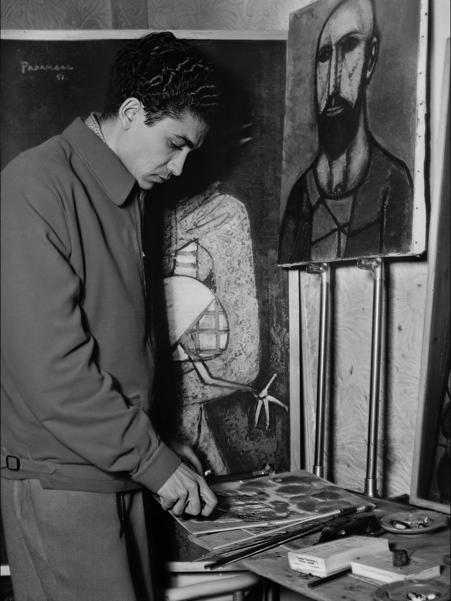 A young man with dark, wavy hair, wearing a jacket, stands at a table looking at a clipboard or piece of paper. Behind him are two paintings—one of a woman with a geometric face and hand, and another of a bearded man with an abstract, elongated face.