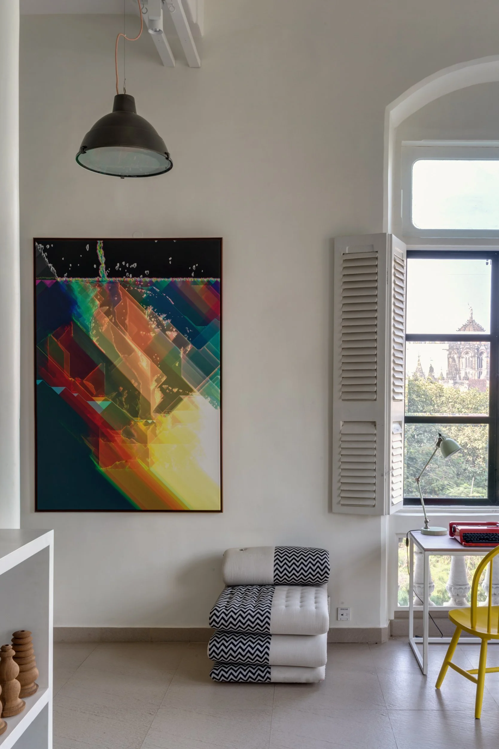 A room with a modern art piece on the wall, a white and black chevron-patterned chair, a small desk with a desk lamp, a yellow chair, a window with view of a historic building, and a black hanging light fixture.