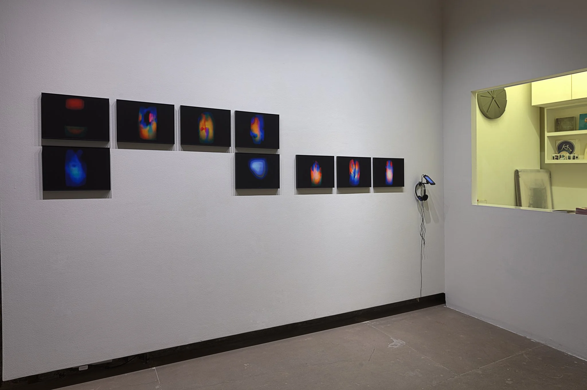 A gallery wall displaying two rows of abstract digital art pieces with colorful heat map-like patterns, mounted on a white wall in a minimalist exhibition space.