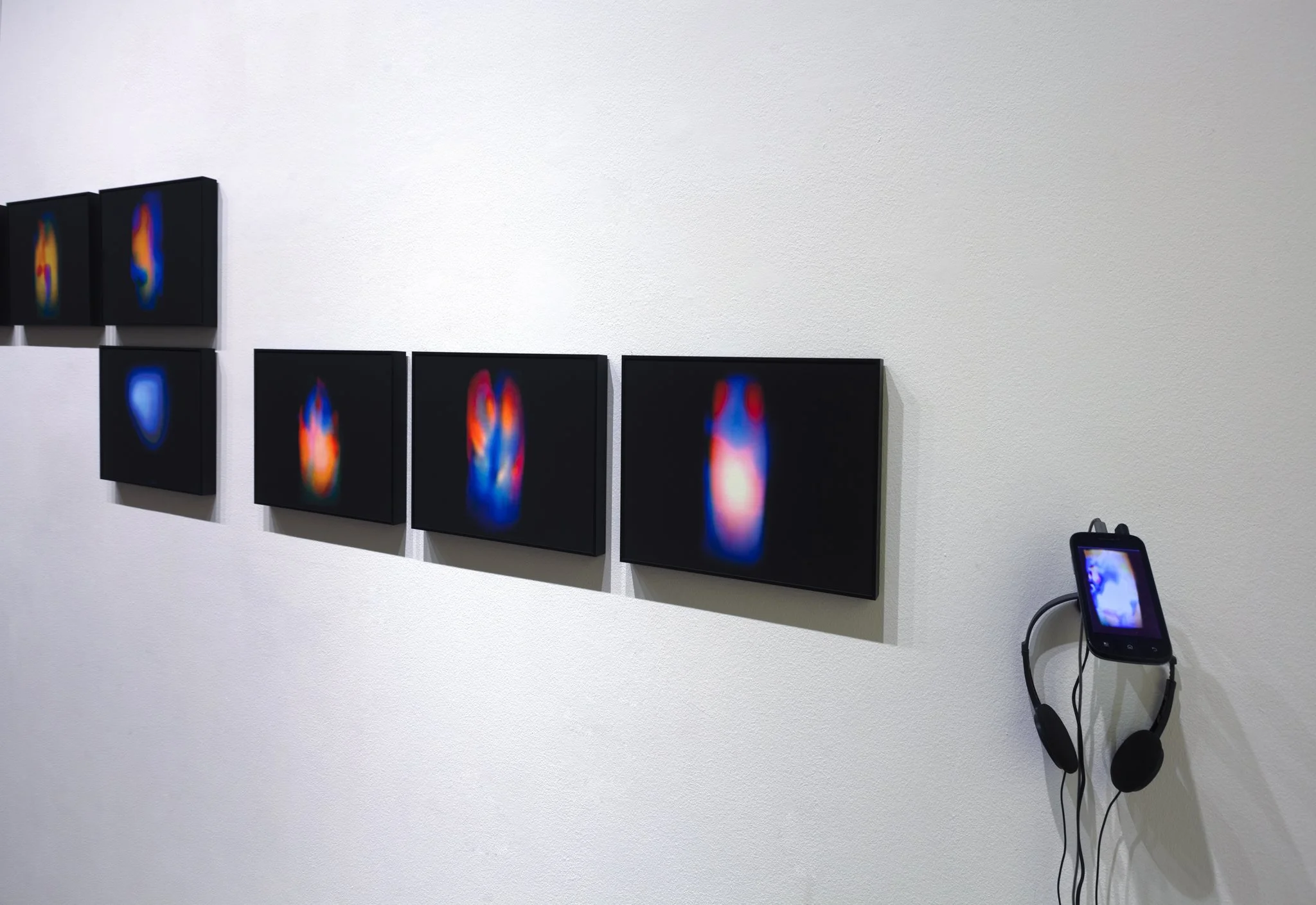 Art gallery display of abstract digital images on black canvases, accompanied by a digital device with headphones hanging on the wall.