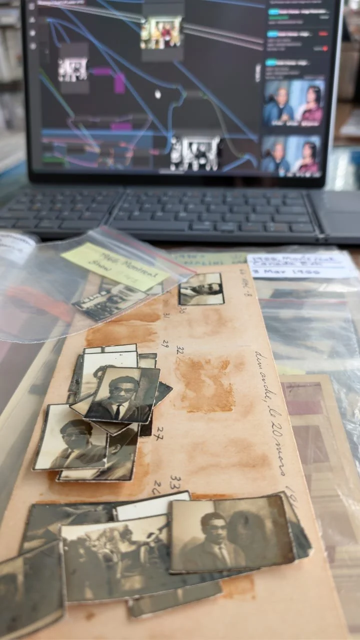 A collection of old black-and-white photographs spread on a table, with a blurred laptop displaying a social media or messaging app in the background.