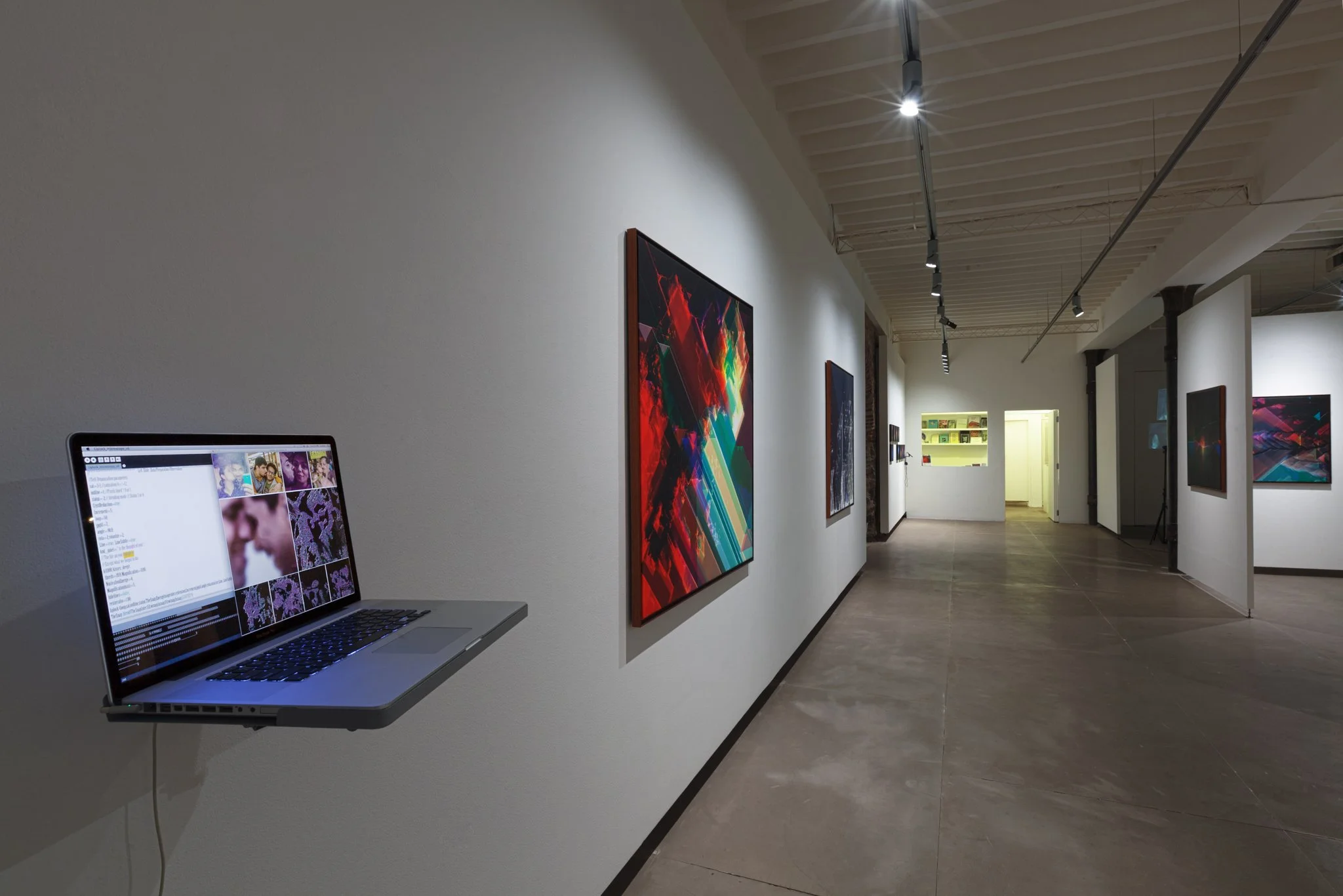 An art gallery with white walls displaying colorful paintings and a laptop mounted on the wall showing a digital image collection.