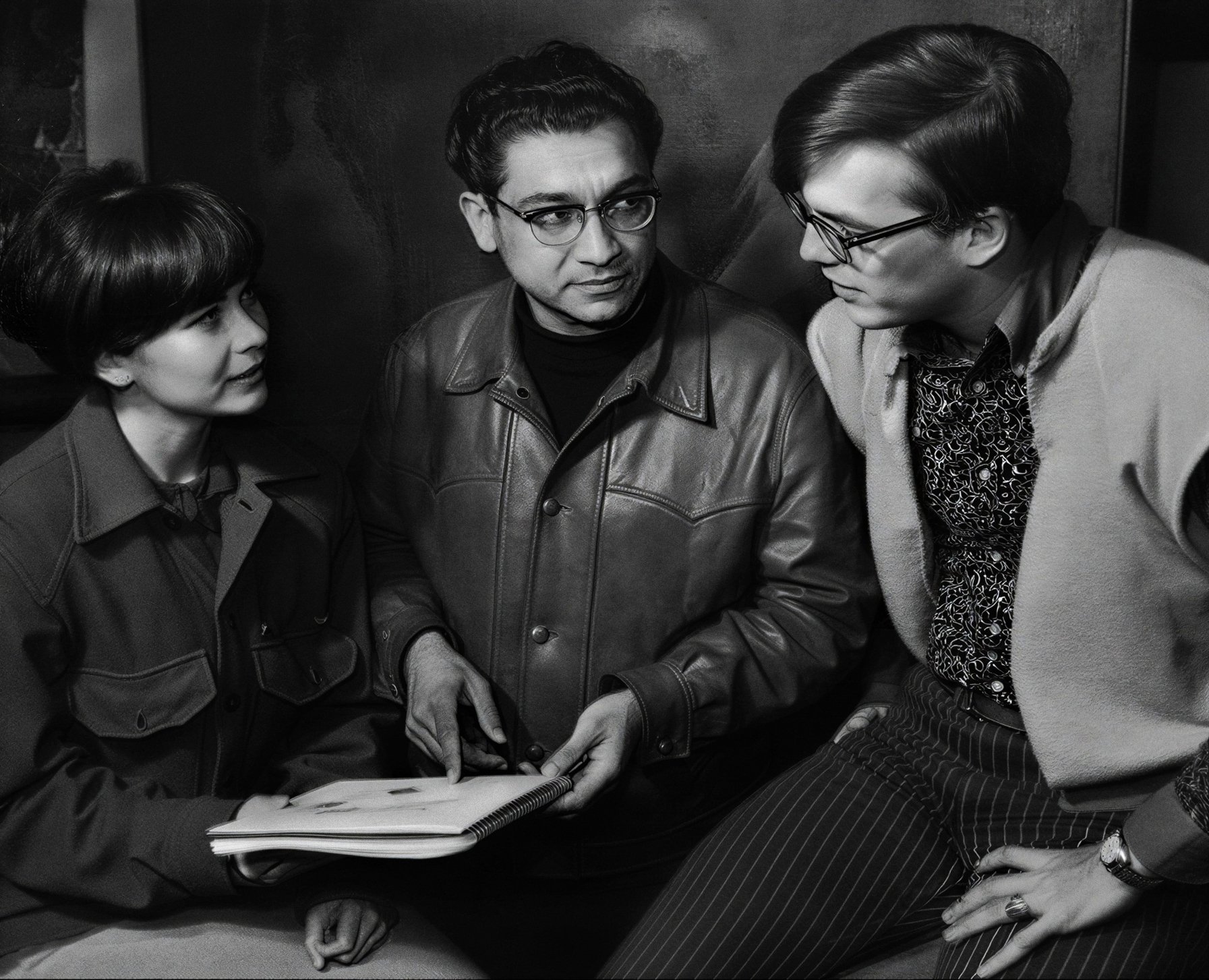 Three people in conversation, two women and one man, with notebooks, black and white photo.