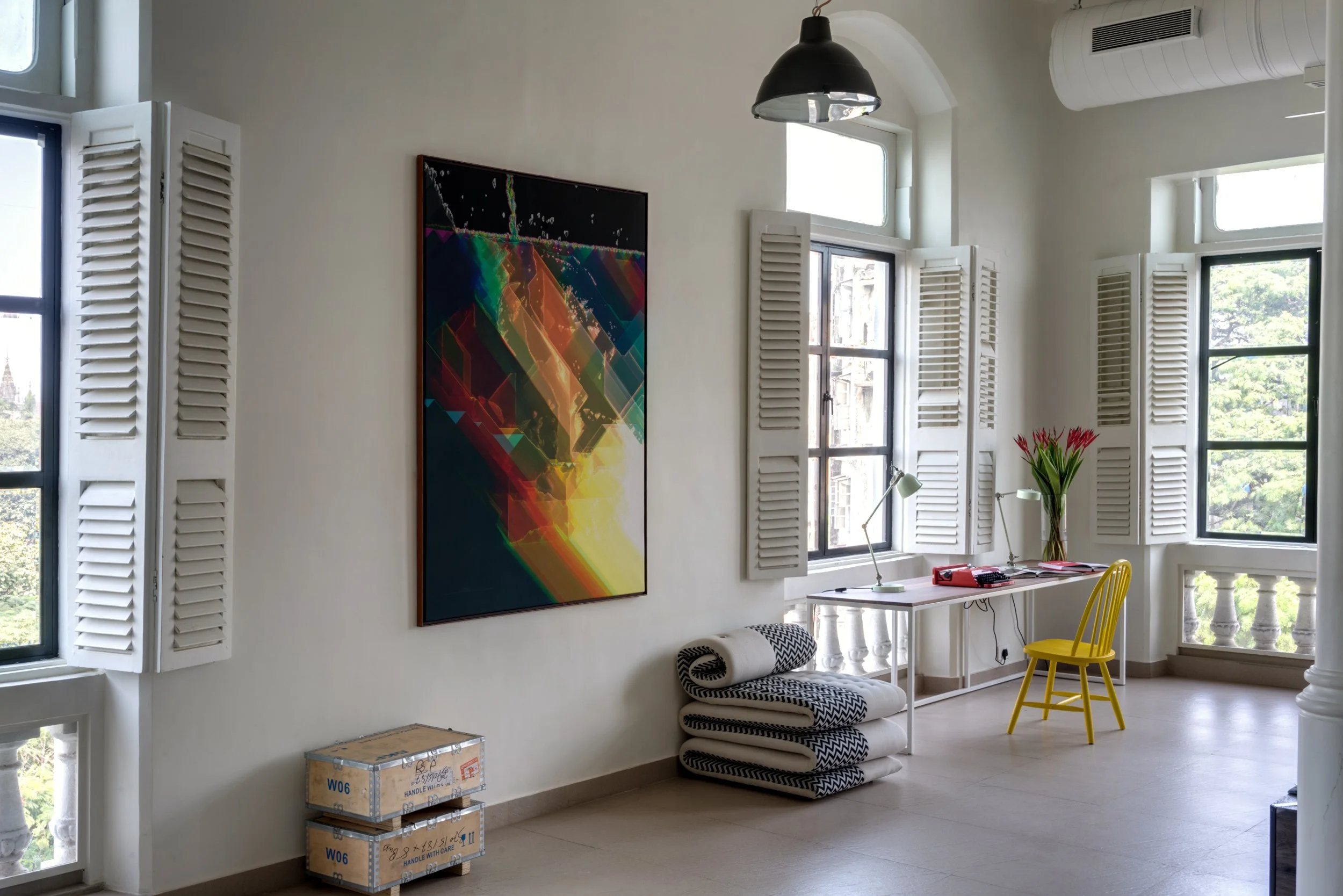 Bright room with white walls and large windows with white shutters, a white desk with a yellow chair, decorative pillows, boxes, a colorful abstract painting on the wall, and a vase of red flowers on the desk.