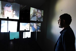 A person looking at multiple video screens on a dark wall, with some screens displaying faces and scenes. Digital Art