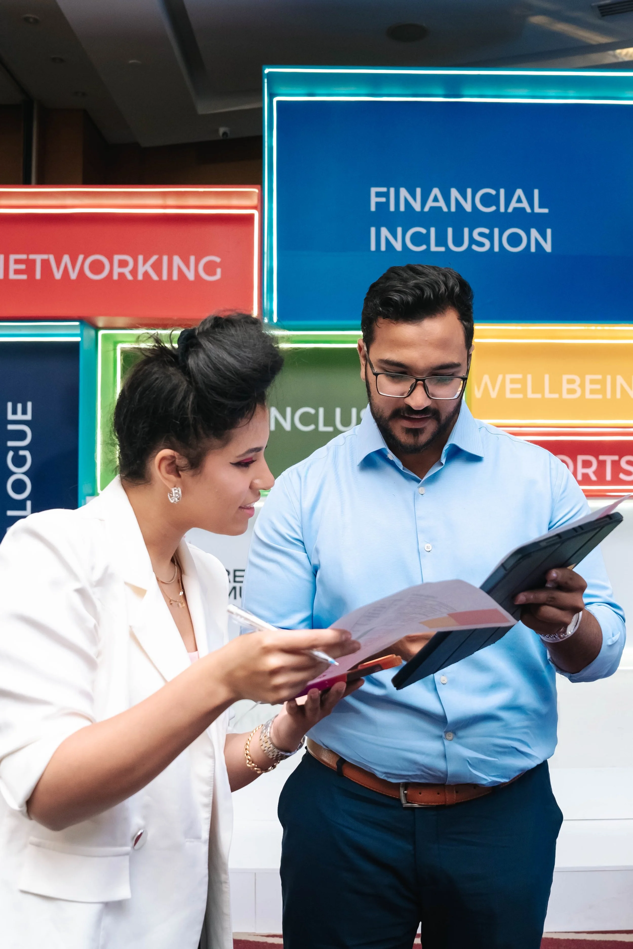 Farah Kordy and another Regional Sustainable Development Forum, First Edition attendee are engaged in a discussion and reviewing documents in front of a colorful background with signs that read 'Networking', 'Financial Inclusion', and 'Wellbeing'.