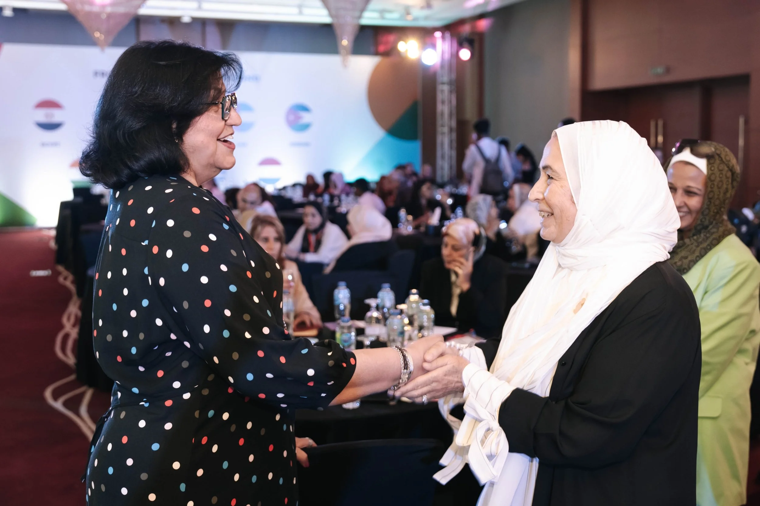 Two women are shaking hands and smiling at each other during Regional Sustainable Development Forum, First Edition. There are people sitting at tables and a large display with logos in the background.