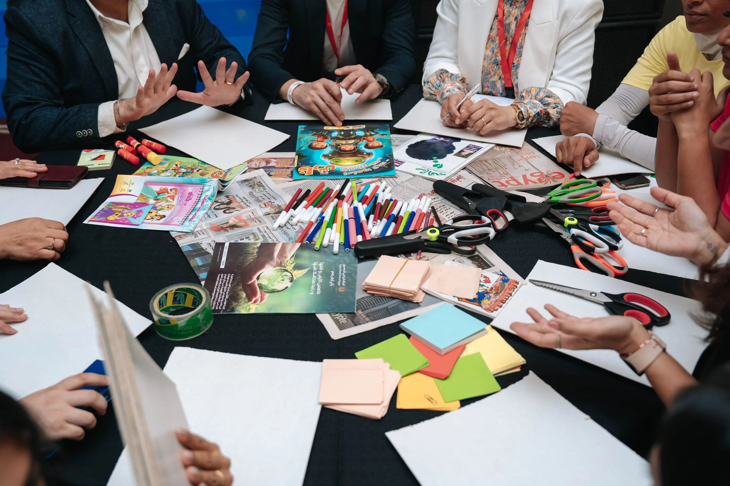 A group of people sitting around a table with arts and crafts supplies at the Regional Sustainable Development Forum, First Edition, including colored markers, scissors, paper, and magazines.