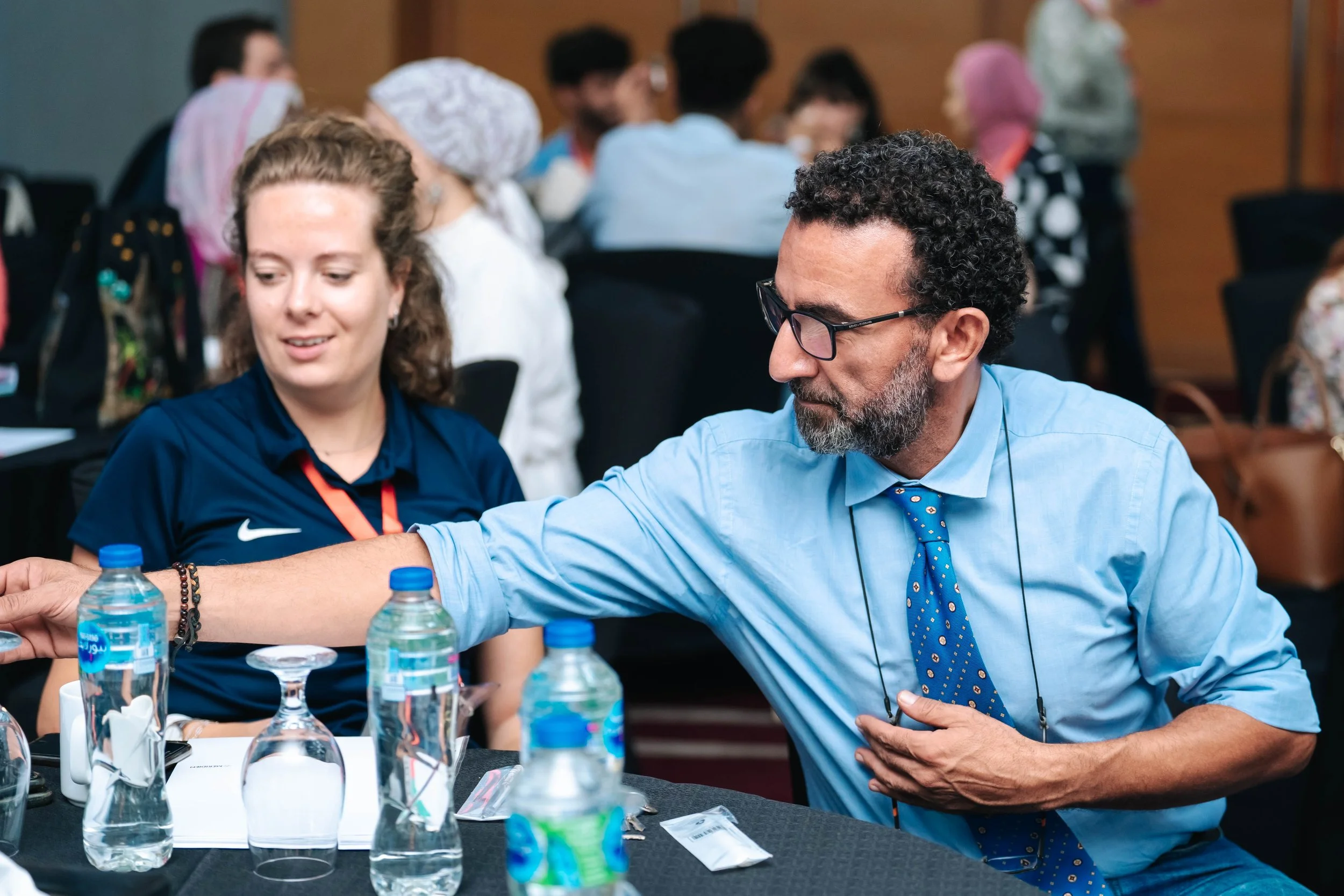 A man reaches across a table filled with water bottles, cups, and paper, while a young woman looks ahead of her; they are in a crowded arena at the Regional Sustainable Development Forum, First Edition.
