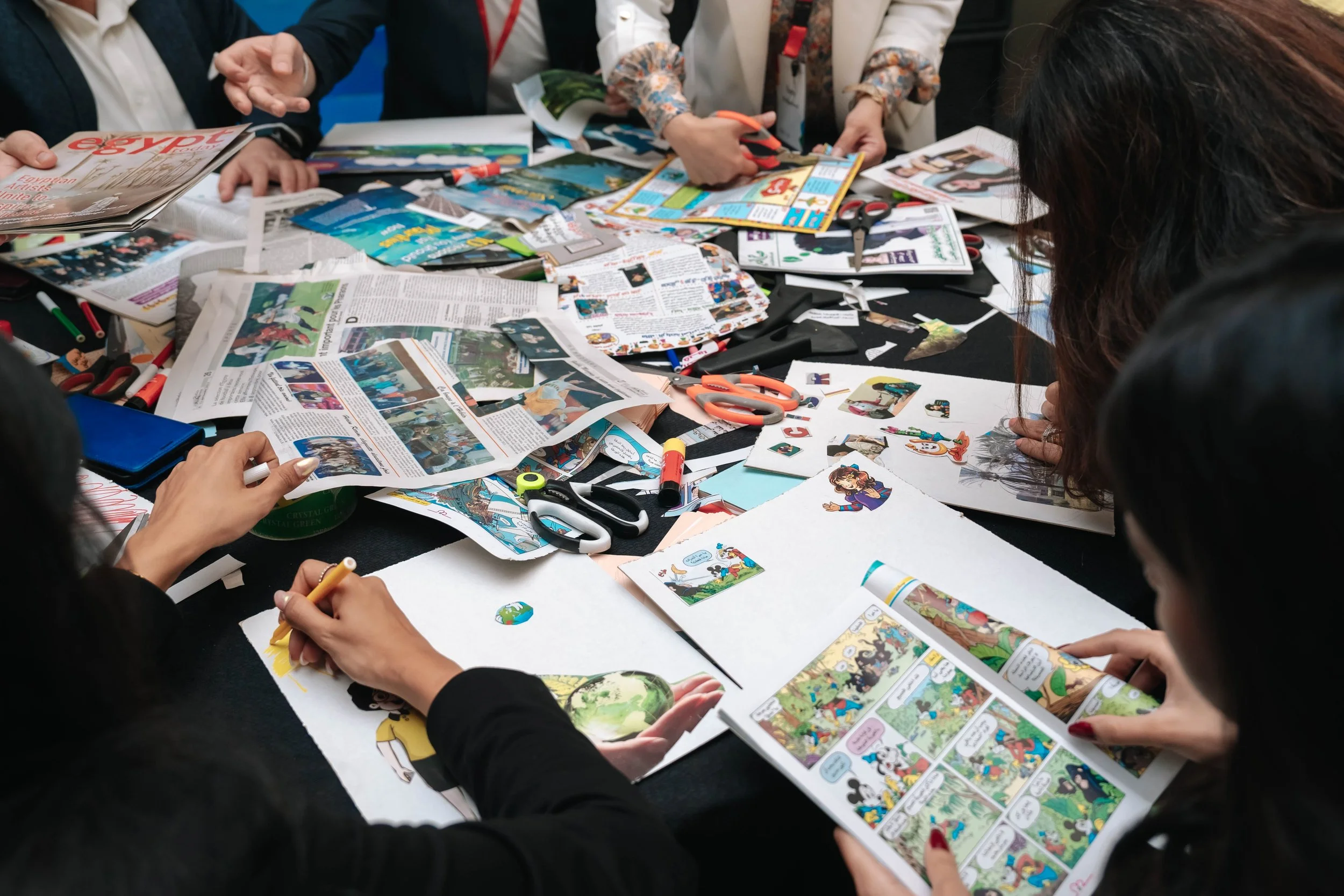 People working on a crafting project at a table covered with various papers, magazines, scissors, markers, and stickers at the Regional Sustainable Development Forum, First Edition