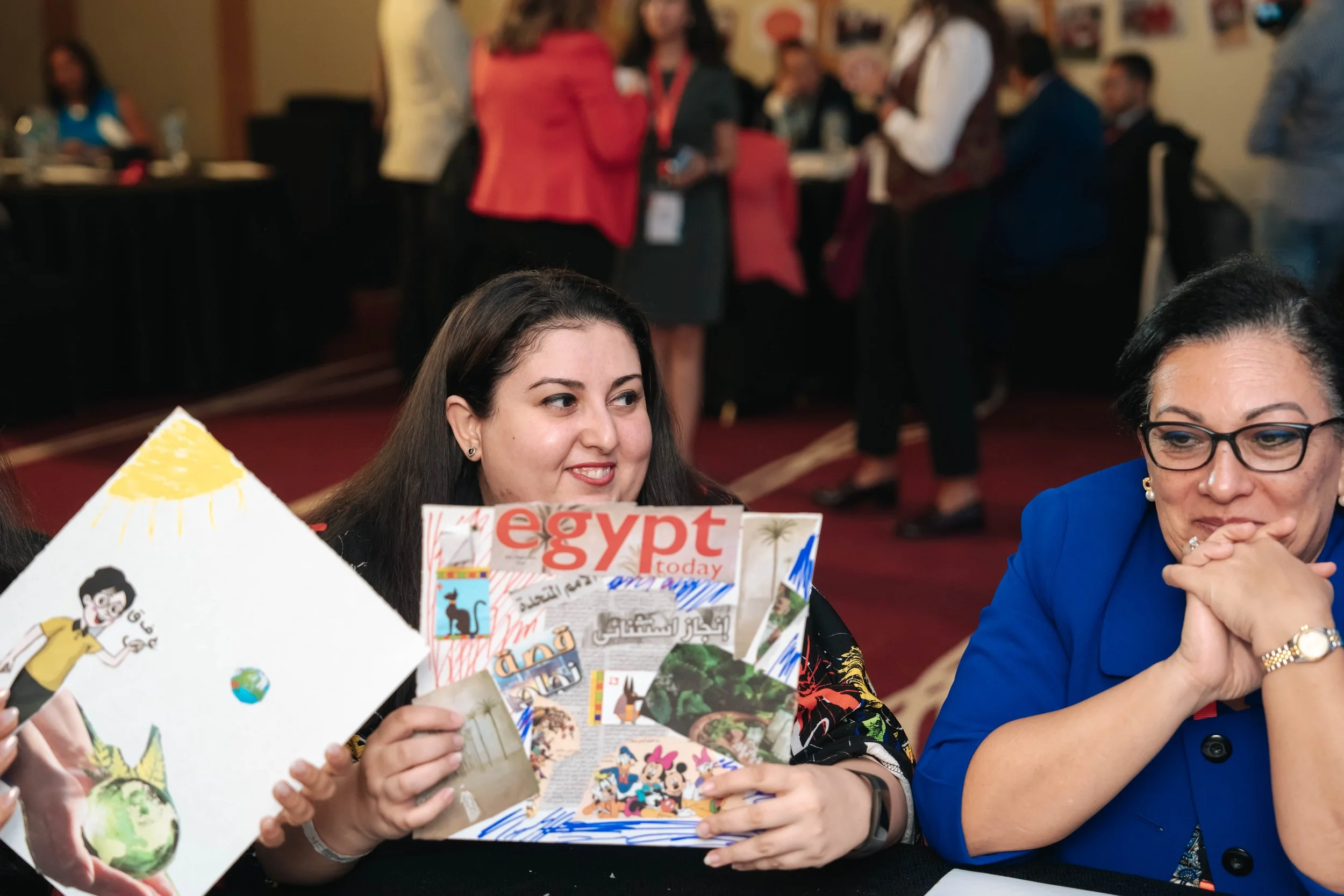 Two women sit at a table, one holding a collage of cutouts including newspapers, photos, and drawings. In the background, people are standing and mingling at the Regional Sustainable Development Forum, First Edition.