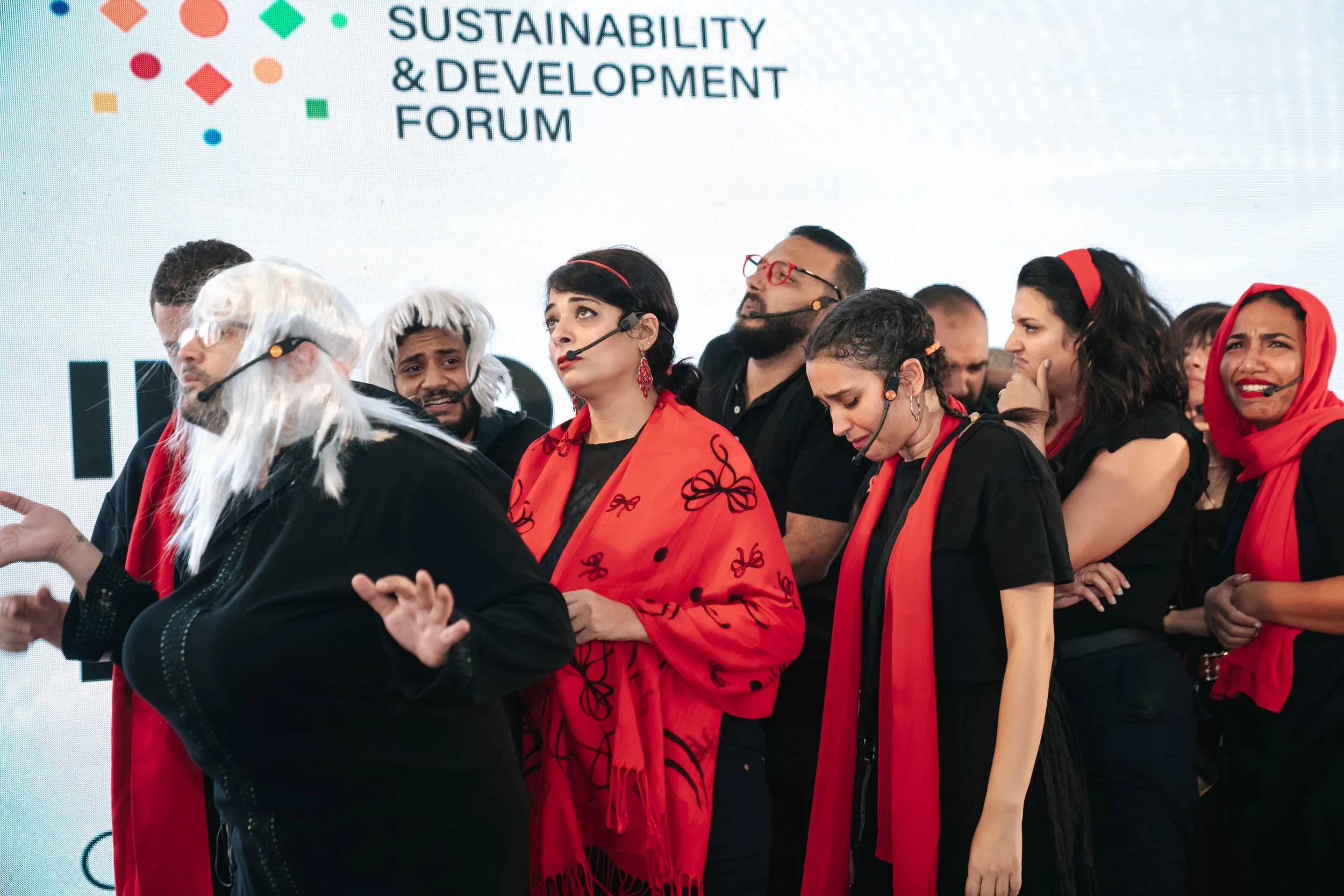 A group of people on stage at the Regional Sustainable Development Forum, First Edition, some wearing black outfits with red scarves or accessories, and a person in a white wig and costume talking with a gesture.