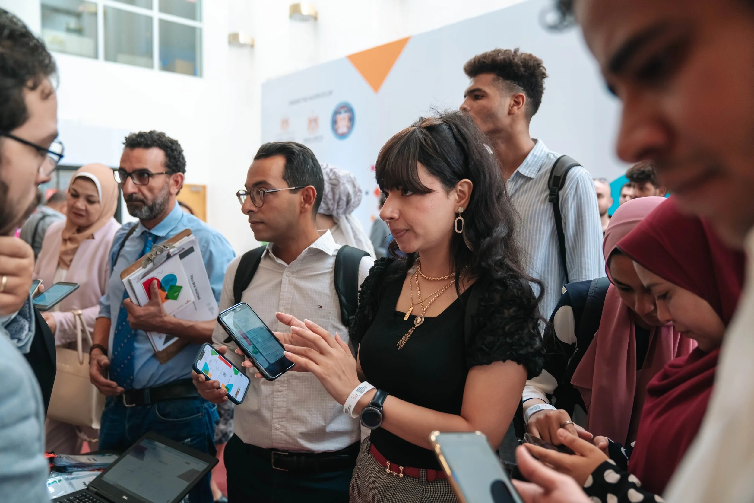 Group of diverse people, including men and women, gathered indoors, looking at their smartphones, at the Regional Sustainable Development Forum, First Edition.