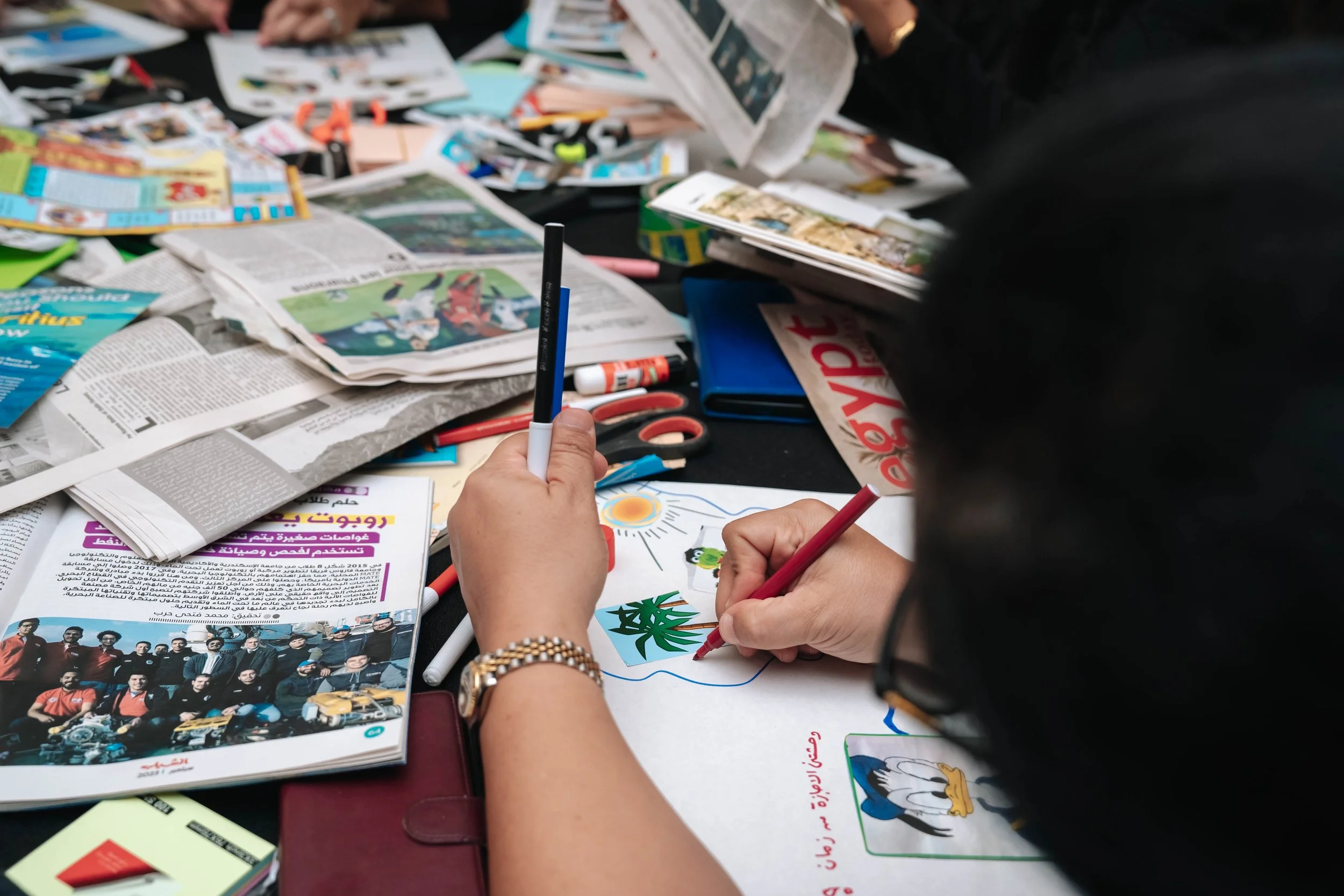 A person coloring a drawing of a palm tree, sun, and water on paper surrounded by magazines, newspapers, scissors, glue, and markers on a black table at a Regional Sustainable Development Forum, First Edition workshop.