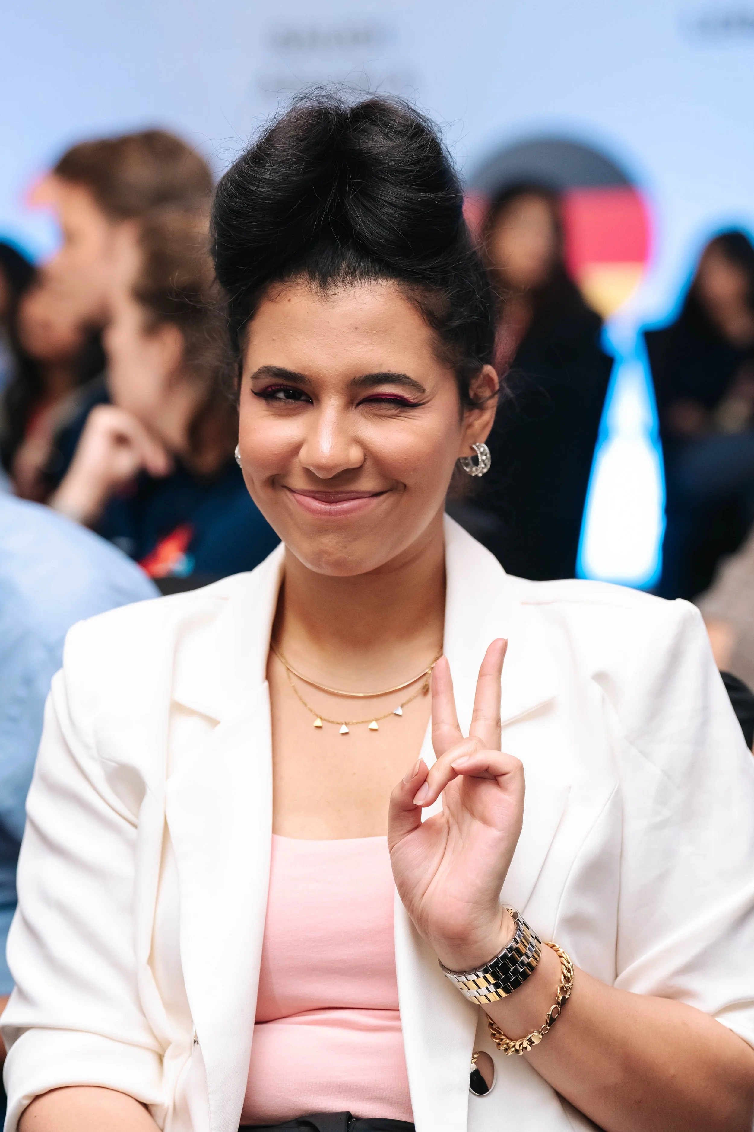 Farah Kordy in white blazer and pink top at the Regional Sustainable Development Forum, First Edition, making a peace sign and winking at the camera, with a blurred crowd in the background.