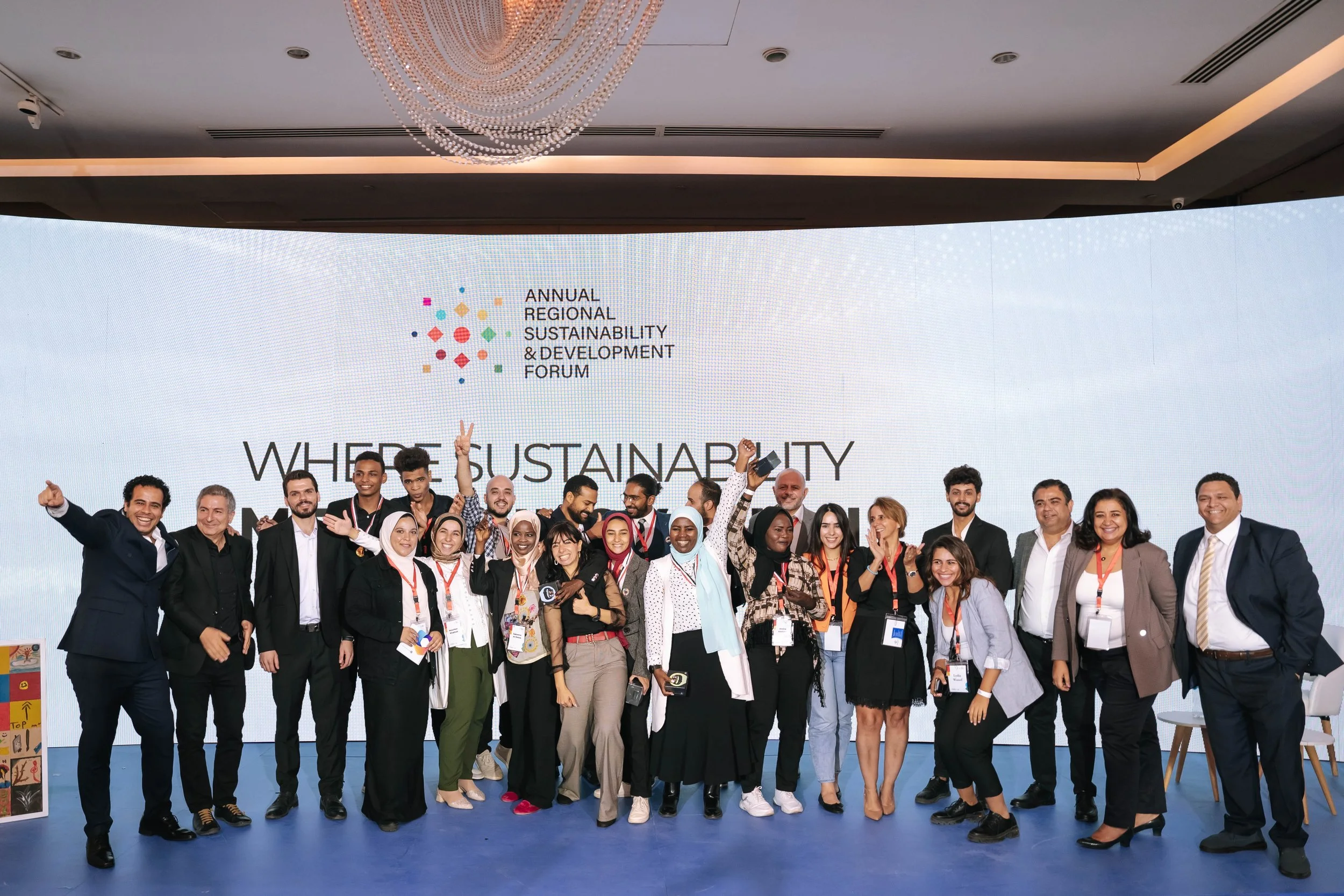 A group photo of all stakeholders at the Regional Sustainable Development Forum, First Edition, smiling, posing for a photo, with a large screen behind them displaying the event's logo and title.