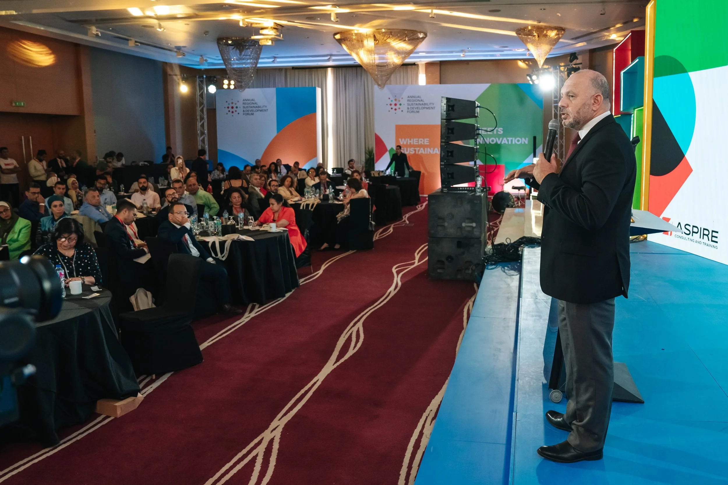 Bassem Emad, CEO of Aspire Consulting International formal giving a keynote speech at RSD Forum, First Edition, with a large audience seated at tables in a hotel conference room.