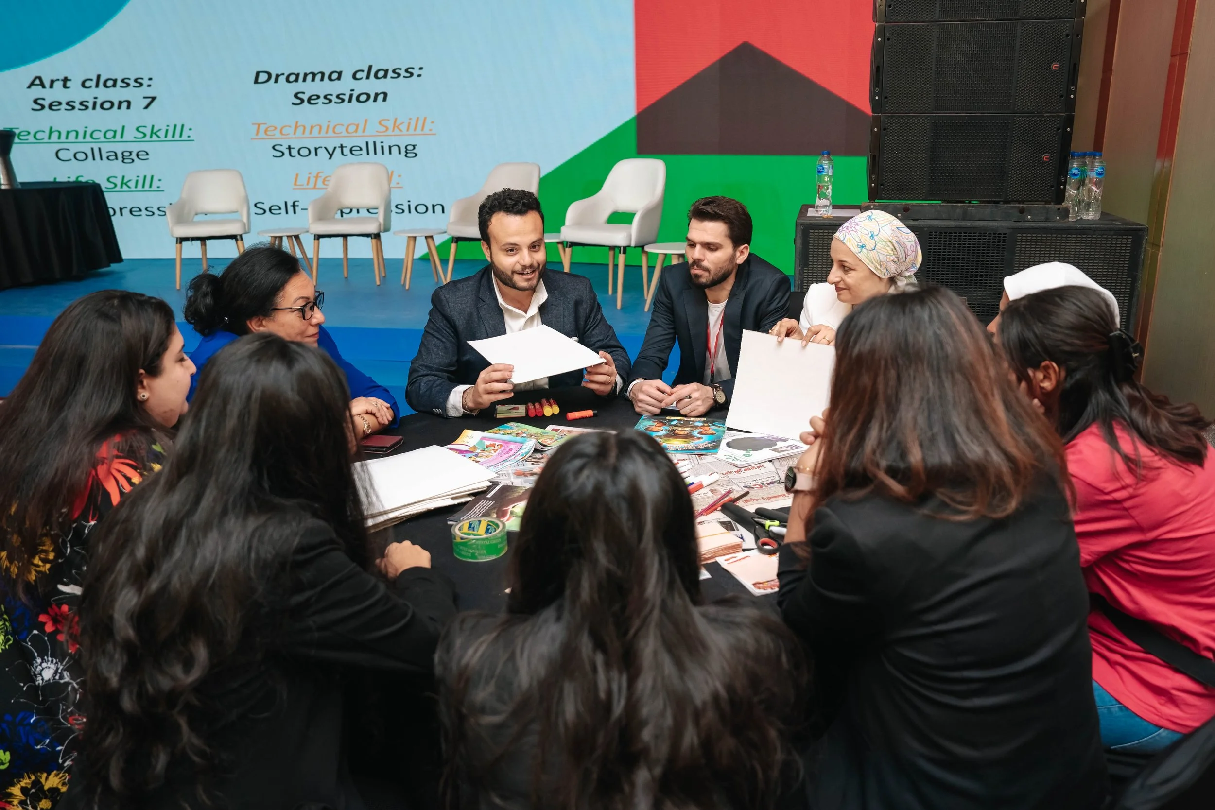Group of people sitting around a table during a workshop oat the Regional Sustainable Development Forum, First Edition, with a presentation screen in the background displaying information about art, drama, and technical skills.