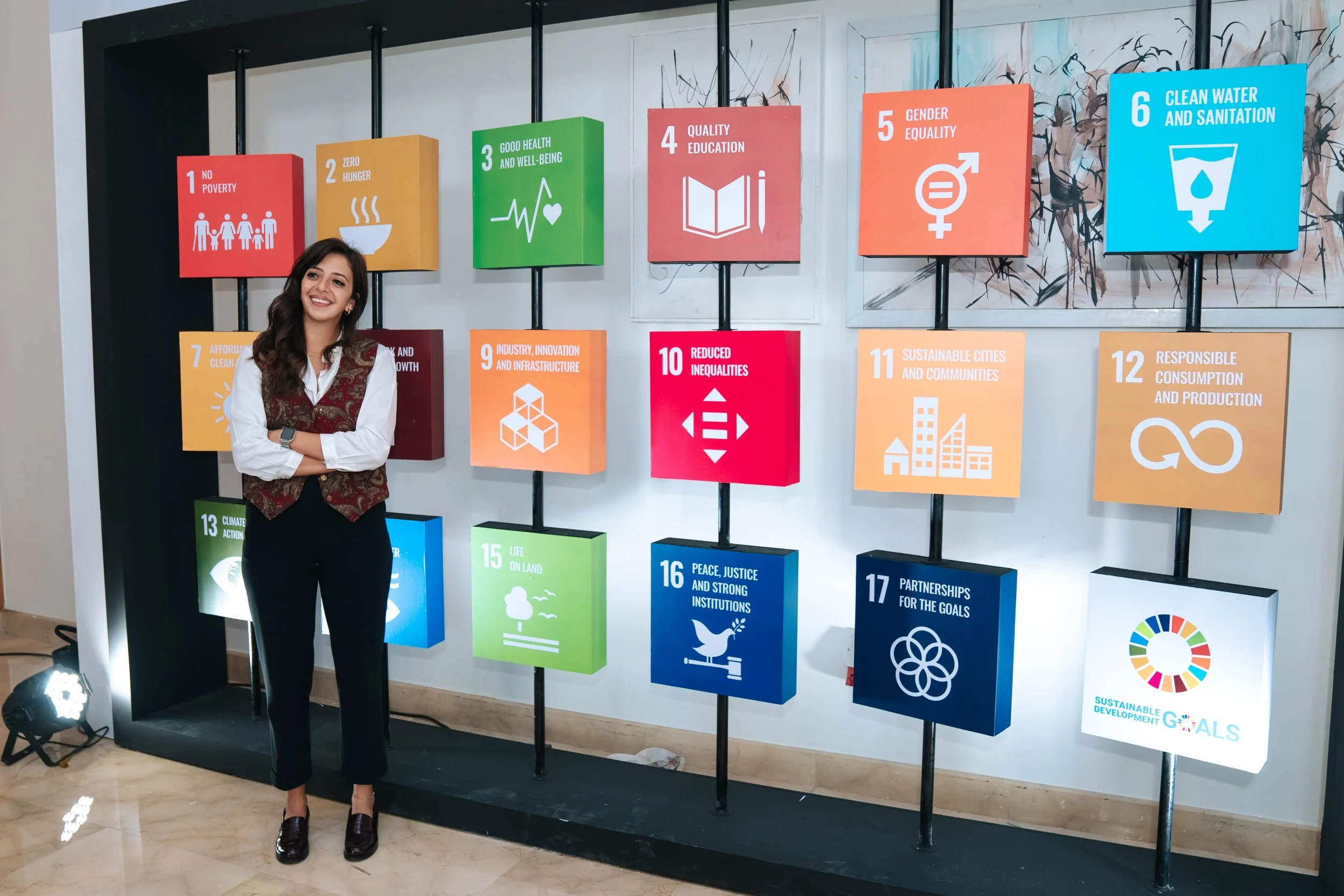 A woman standing and smiling in front of a display of colorful icons representing the United Nations Sustainable Development Goals on a white wall with the overarching Regional Sustainable Development Forum logo.