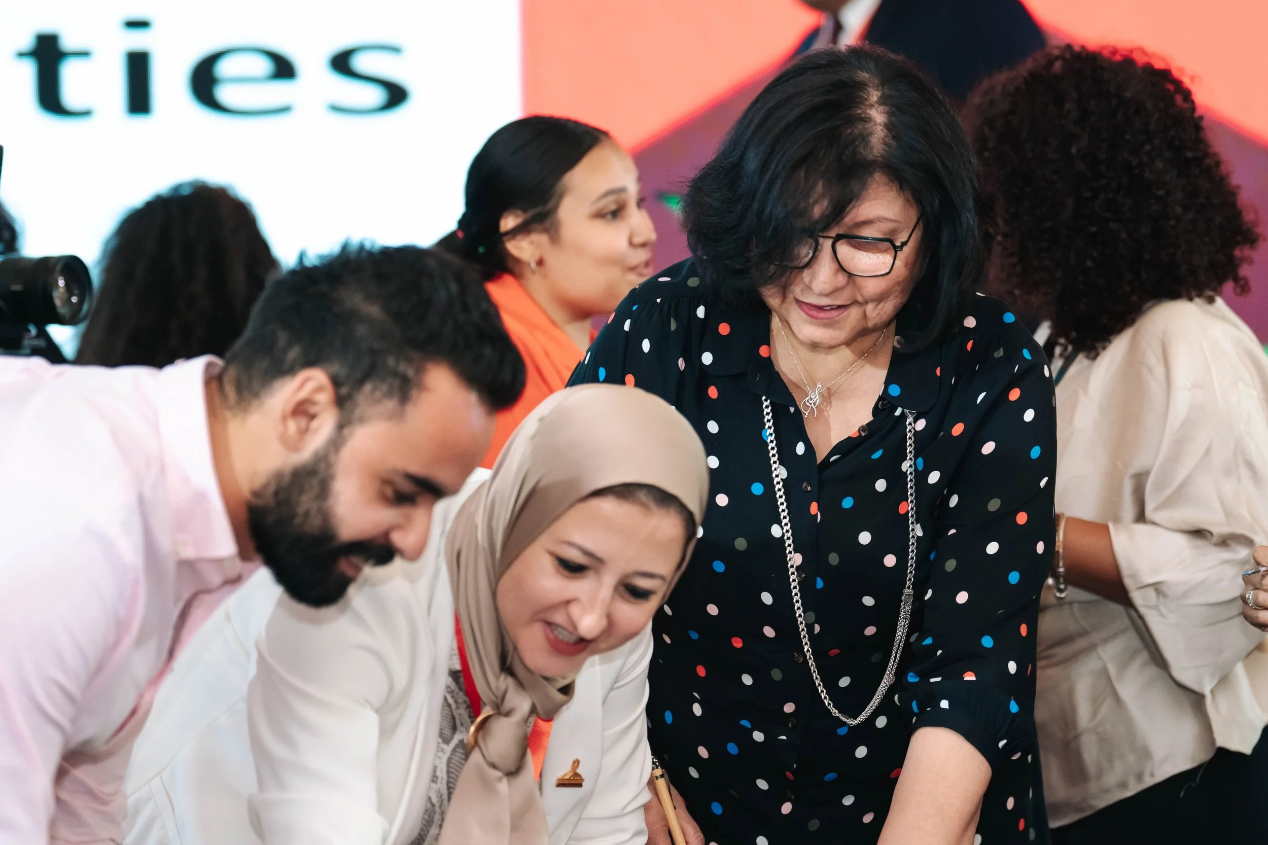 Several diverse women smiling and engaging in a discussion at Regional Sustainable Development Forum, First Edition, with a large screen displaying partially visible text in the background.