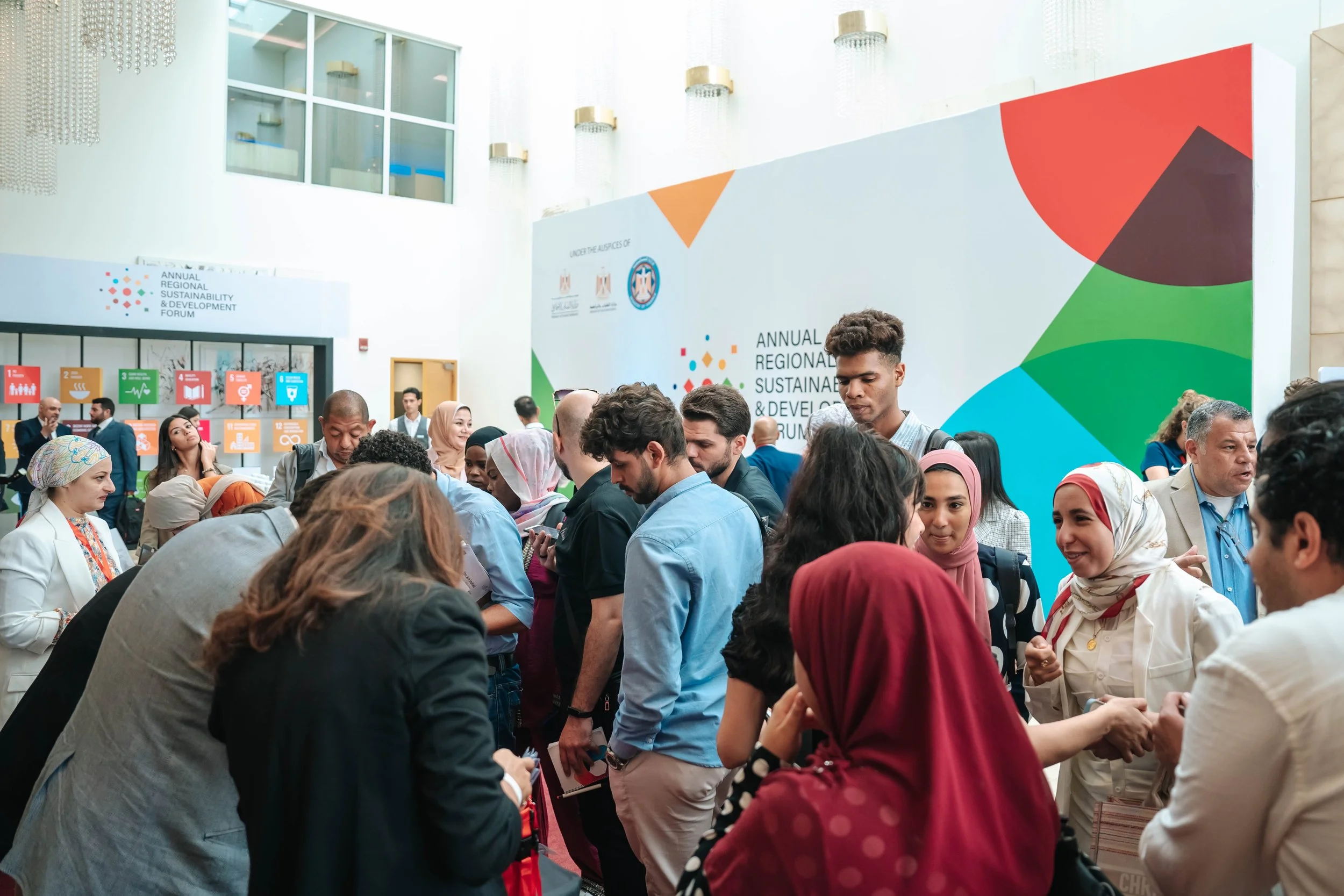 A diverse group of people gathering and networking at the Regional Sustainable Development Forum, First Edition, with a large colorful banner in the background and display panels featuring United Nations Sustainable Development Goals.