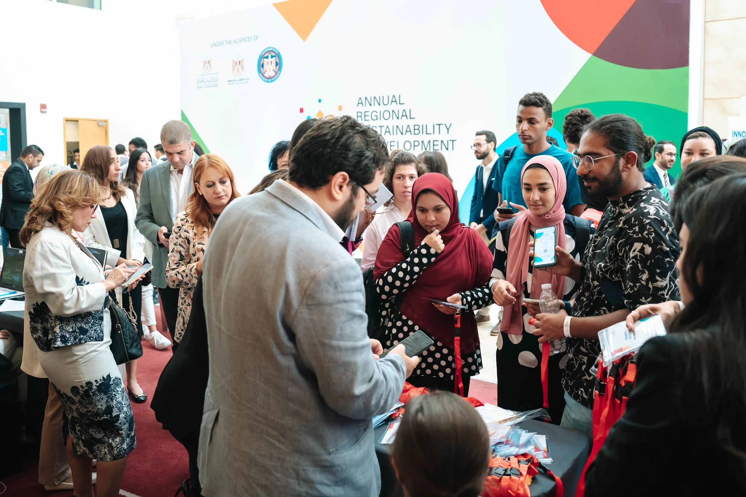 A diverse group of people gathered around a conference table at the Regional Sustainable Development Forum, First Edition, engaging with various information materials and devices.