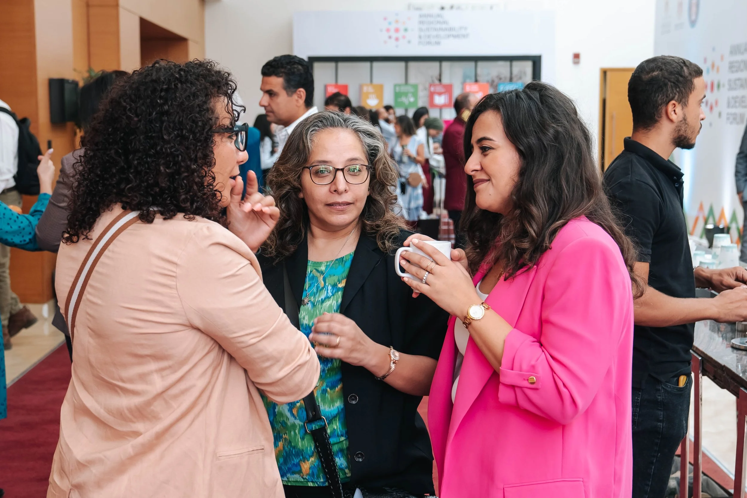 Three women engaging in conversation at the Regional Sustainable Development Forum, First Edition, with other attendees in the background.