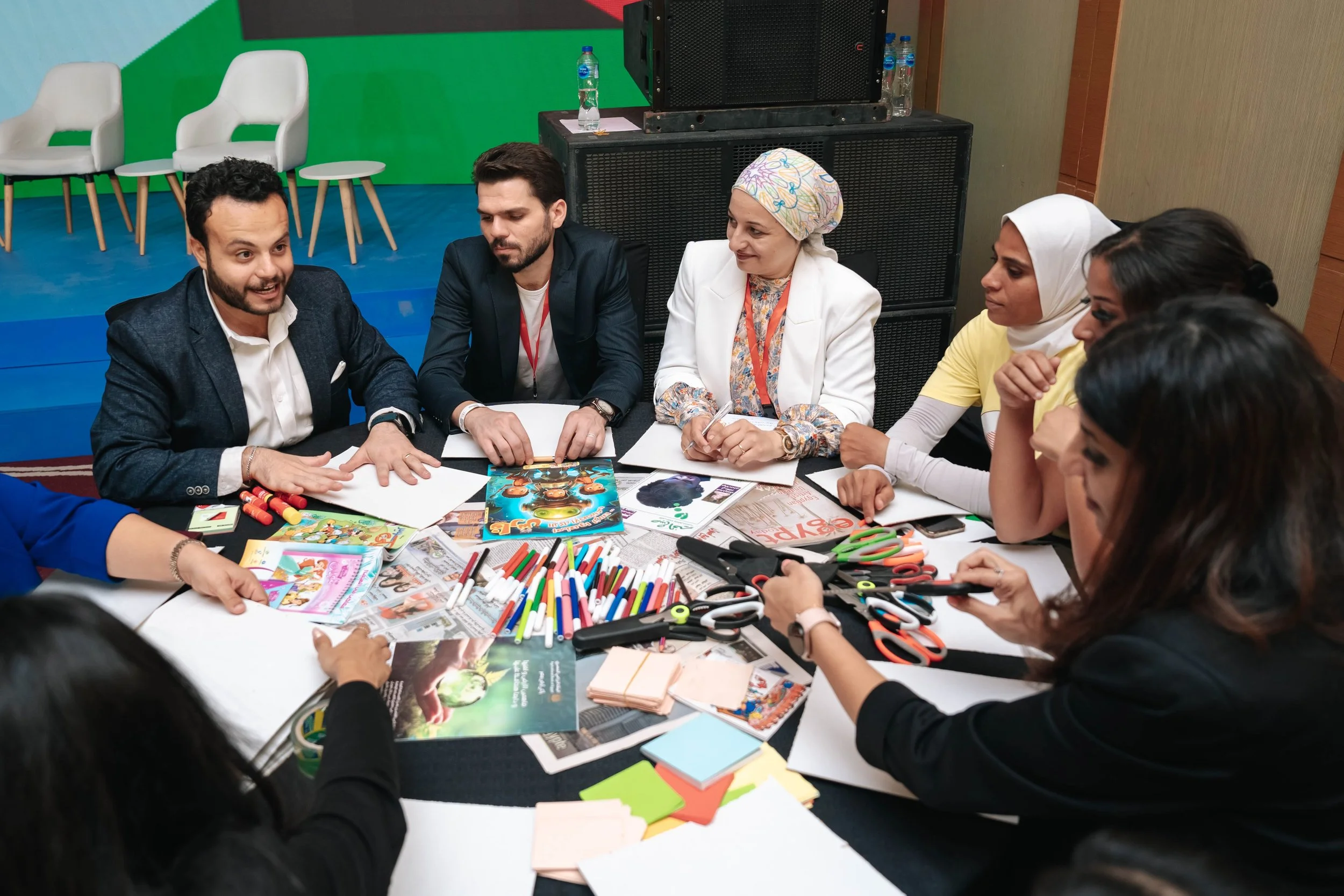 A diverse group of women and men engaging in a meeting or workshop at the Regional Sustainable Development Forum, First Edition, all sitting around a table filled with arts and craft supplies, including markers, scissors, and magazines.