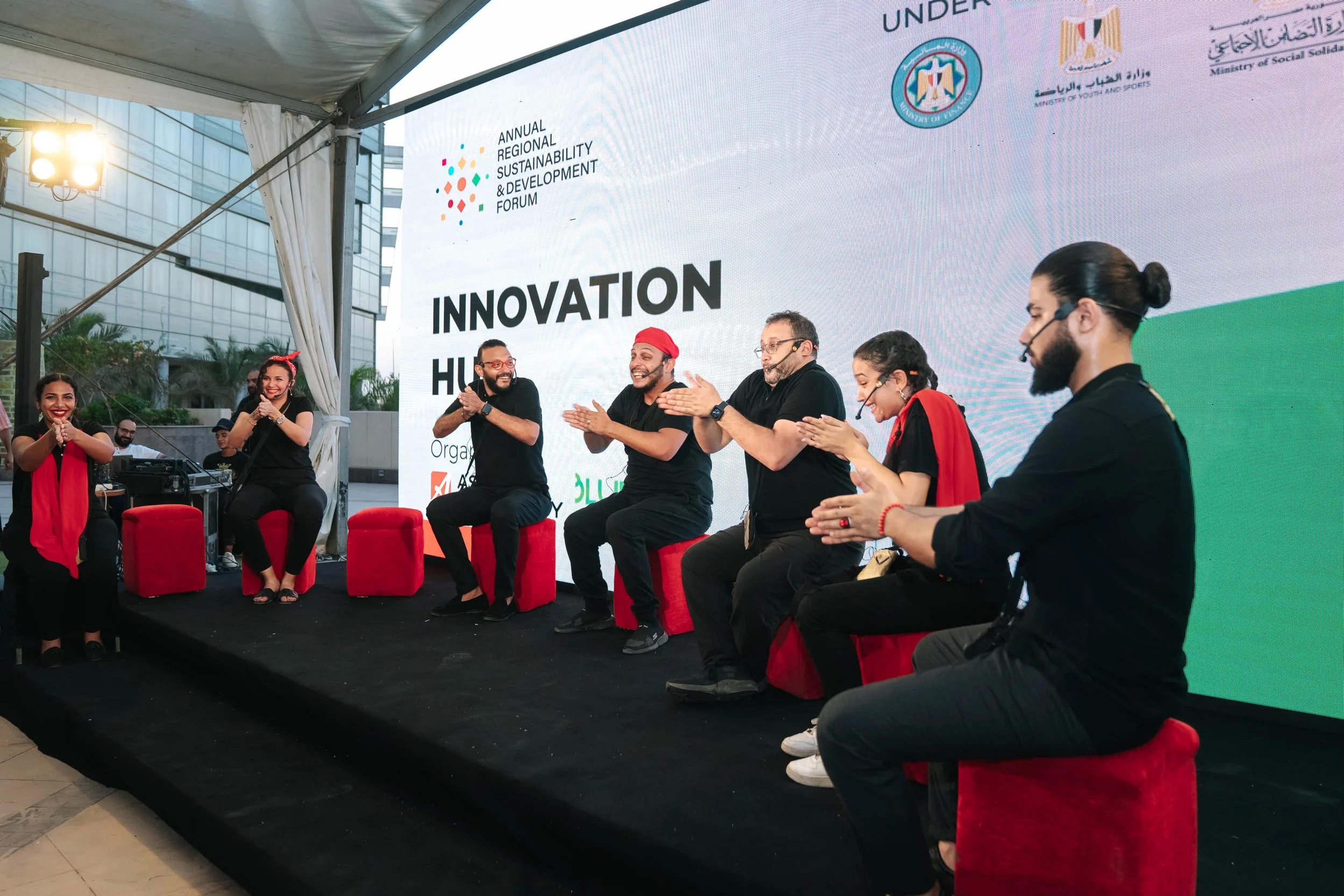 A group of people on stage playing instruments at the Regional Sustainable Development Forum, First Edition, with a large digital screen behind them displaying event details and logos, some of the panelists are clapping and smiling.