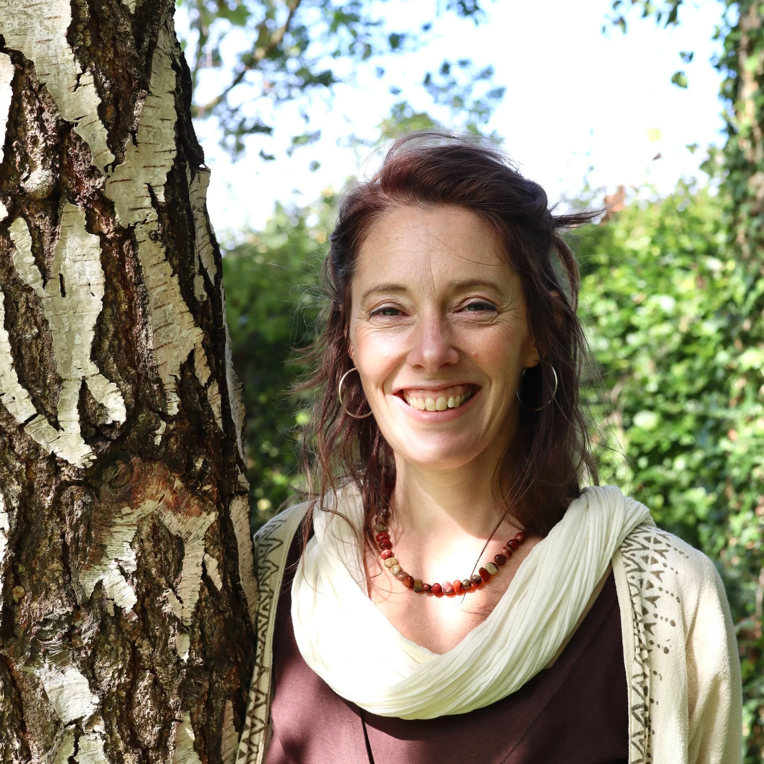 A woman with shoulder-length brown hair standing outdoors next to a tree, smiling with visible teeth, wearing hoop earrings, a beaded necklace, a cream-colored scarf, and a brown top, with green foliage and blue sky in the background.