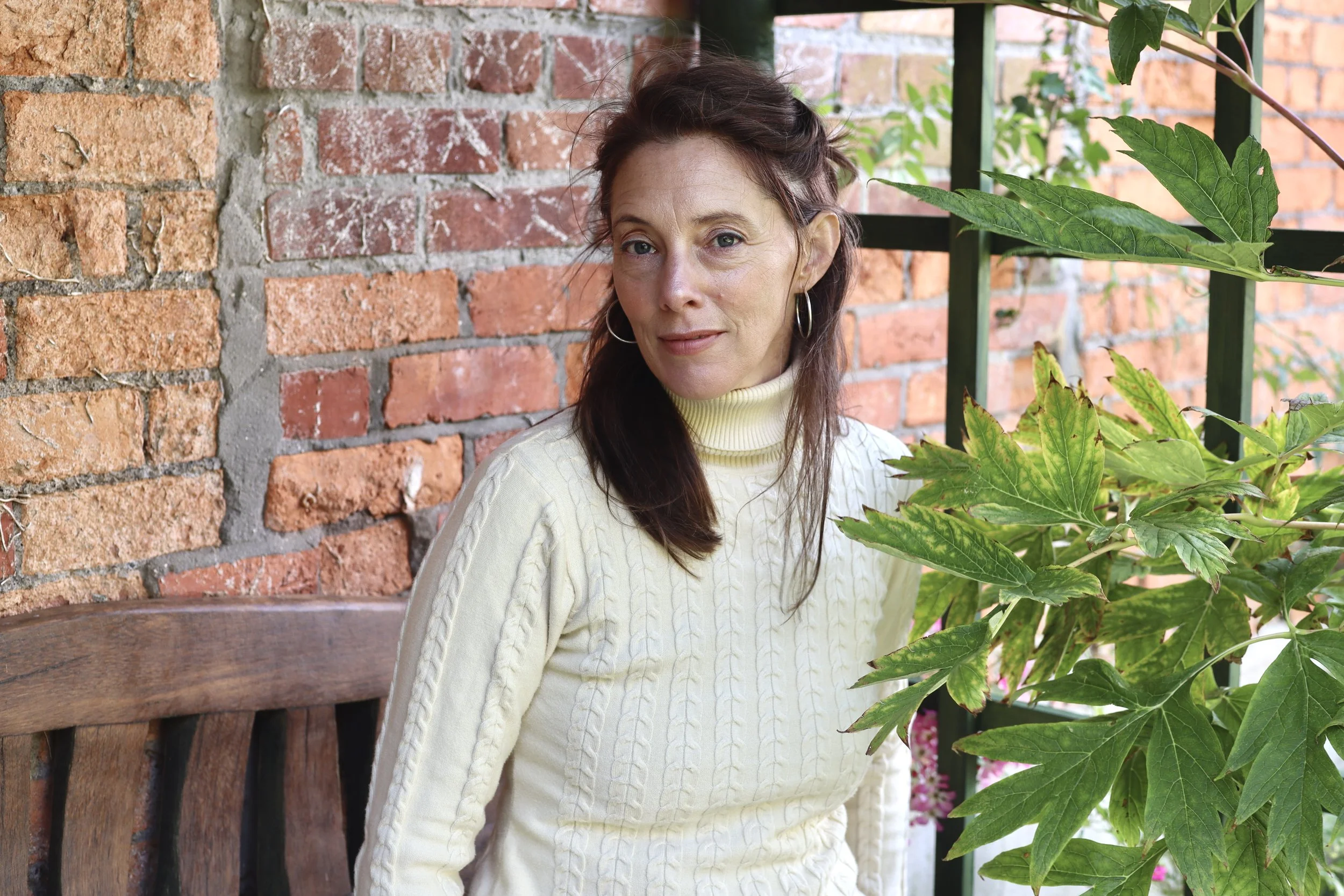 A woman with earrings and brown hair, wearing a light yellow turtleneck sweater, sitting on a wooden bench next to a large leafy plant, with a brick wall and greenery in the background.
